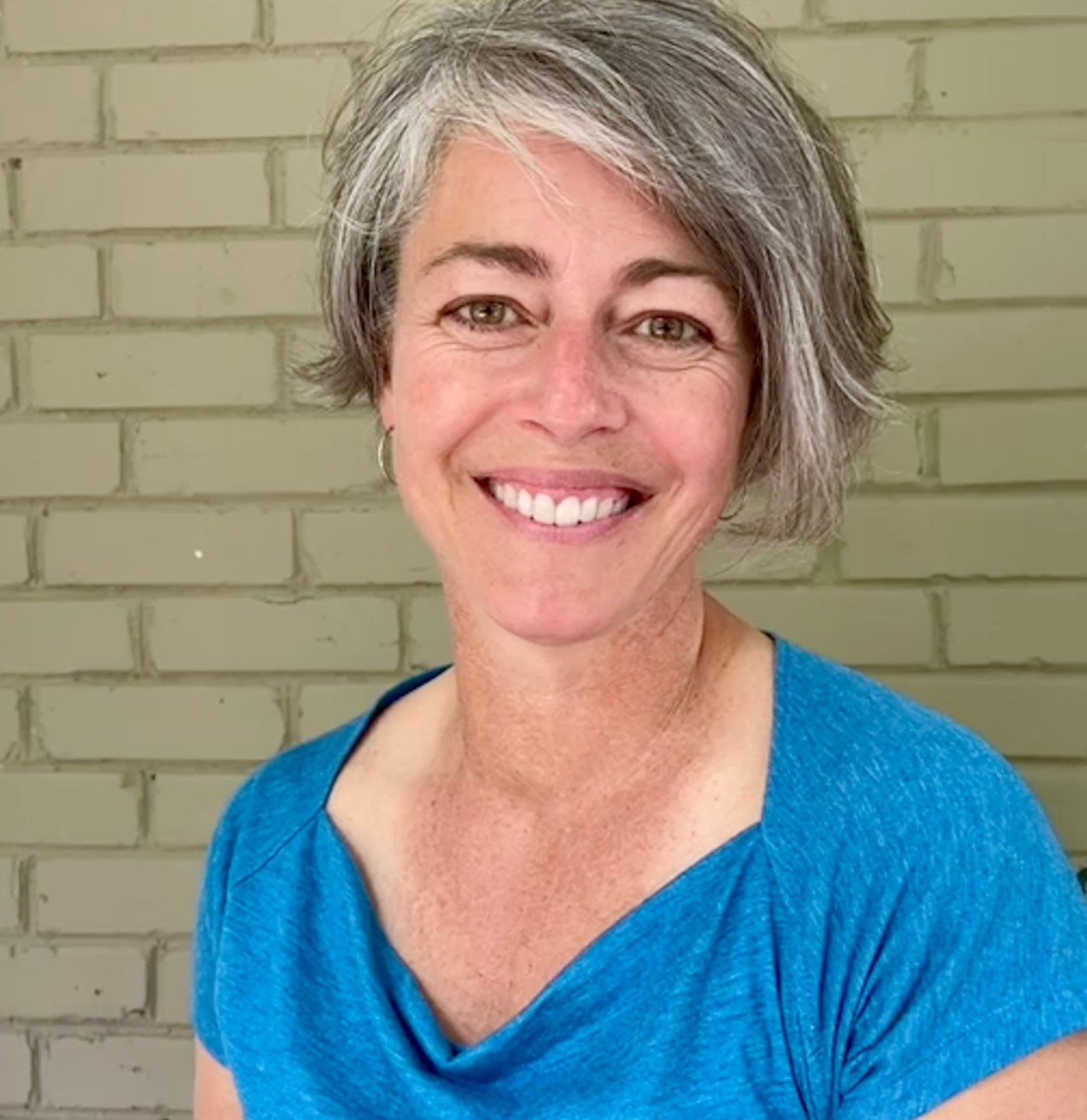 A certified hypnotherapist who used the spiritual hypnosis preparation kit for past life regression and Life Between Lives Hypnotherapy in a blue shirt smiling against a brick wall.