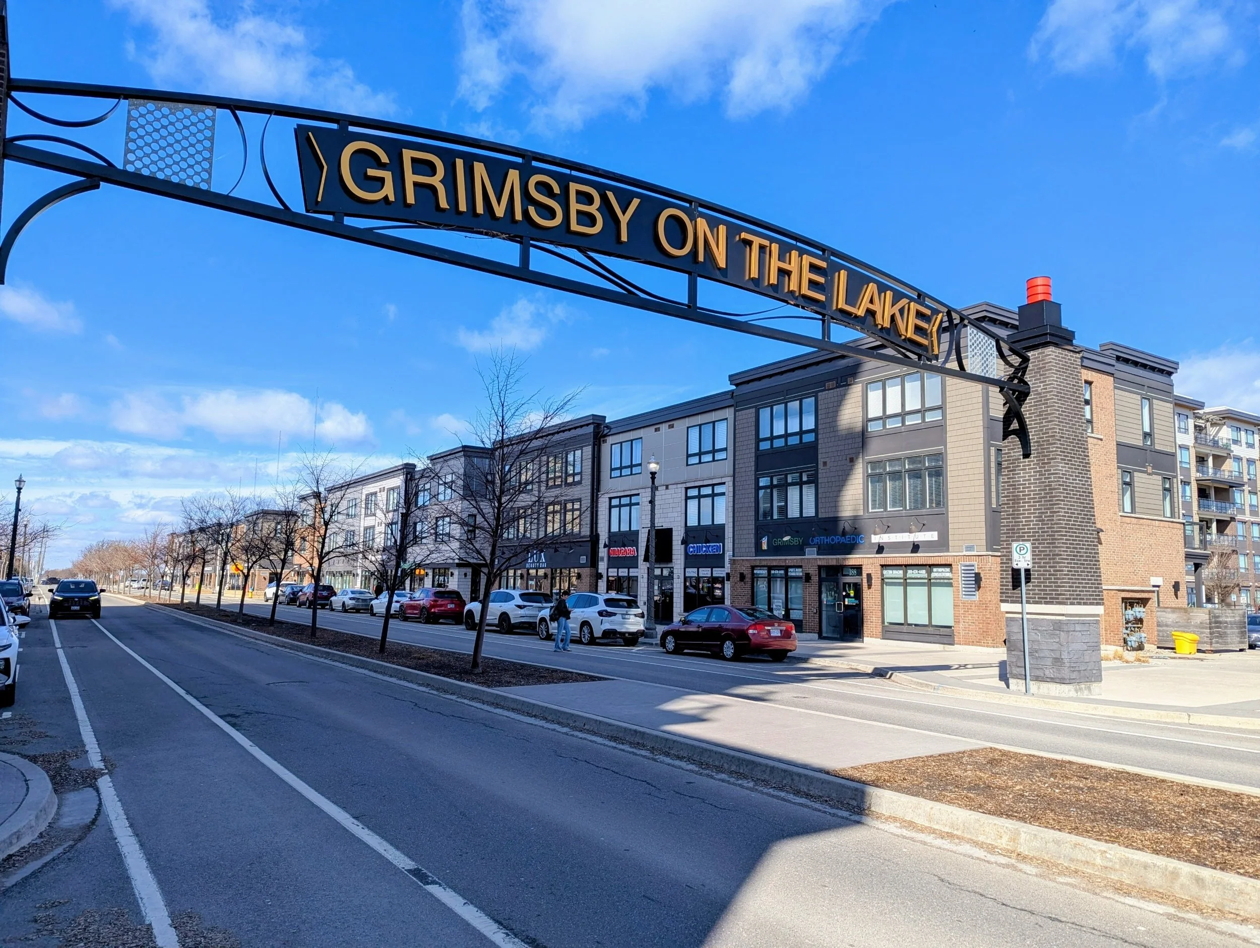 A street scene in a commercial area with modern buildings facing the sidewalk, parked cars along the street, and a large sign overhead that reads 'Grimsby on the Lake' against a blue sky with some clouds.
