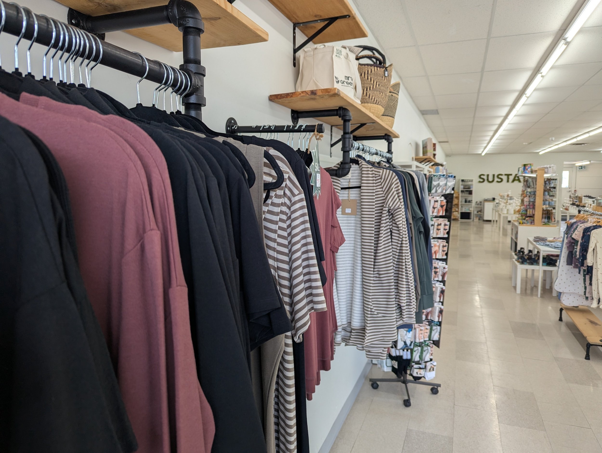 Clothing display in a retail store with shirts on black racks, shelves with baskets and bags, and store signage visible in the background.