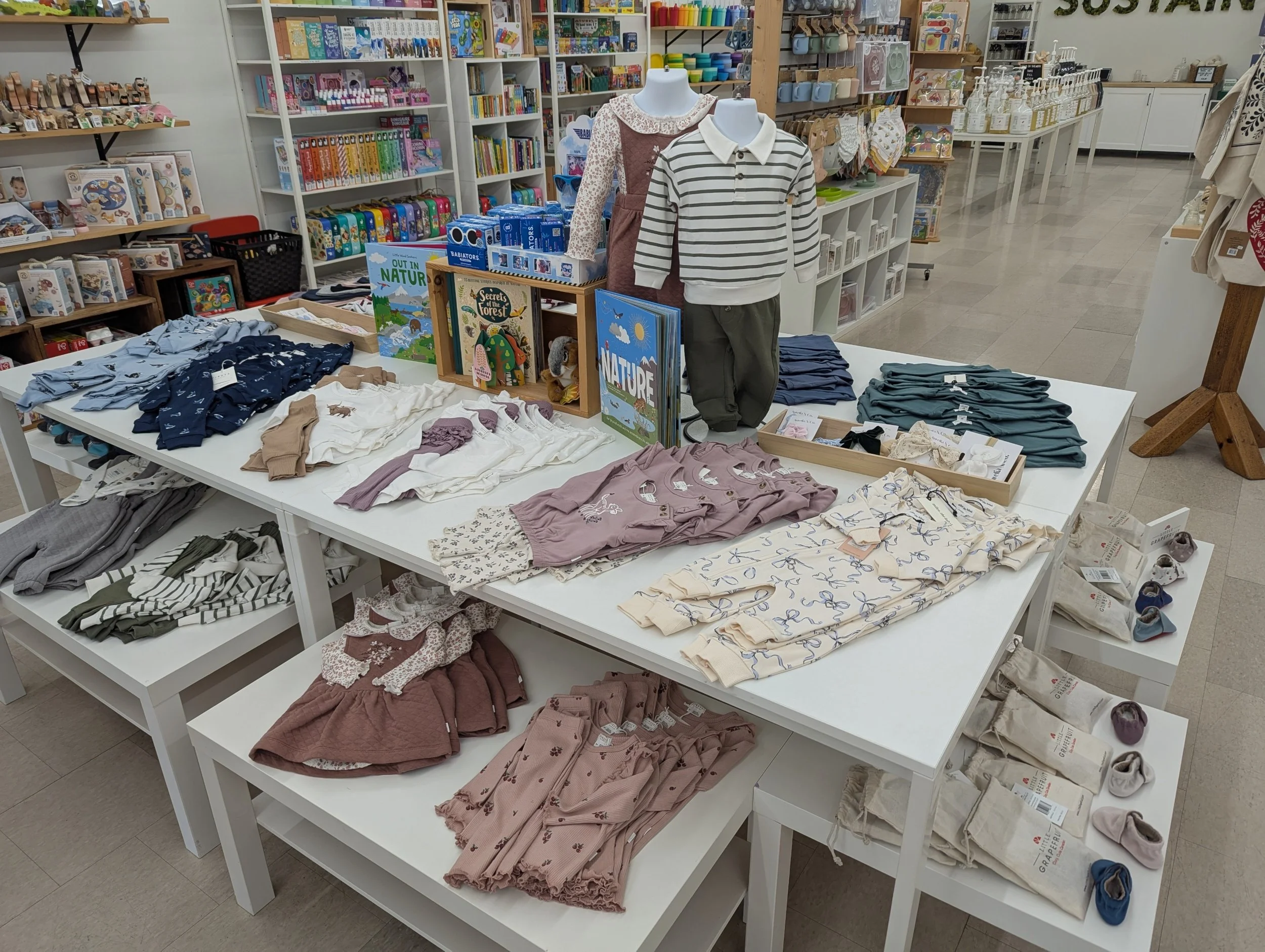 Children's clothing display table with folded shorts and socks, mannequins wearing long-sleeve shirts and pants, surrounding shelves filled with books, toys, and household items in a retail store.