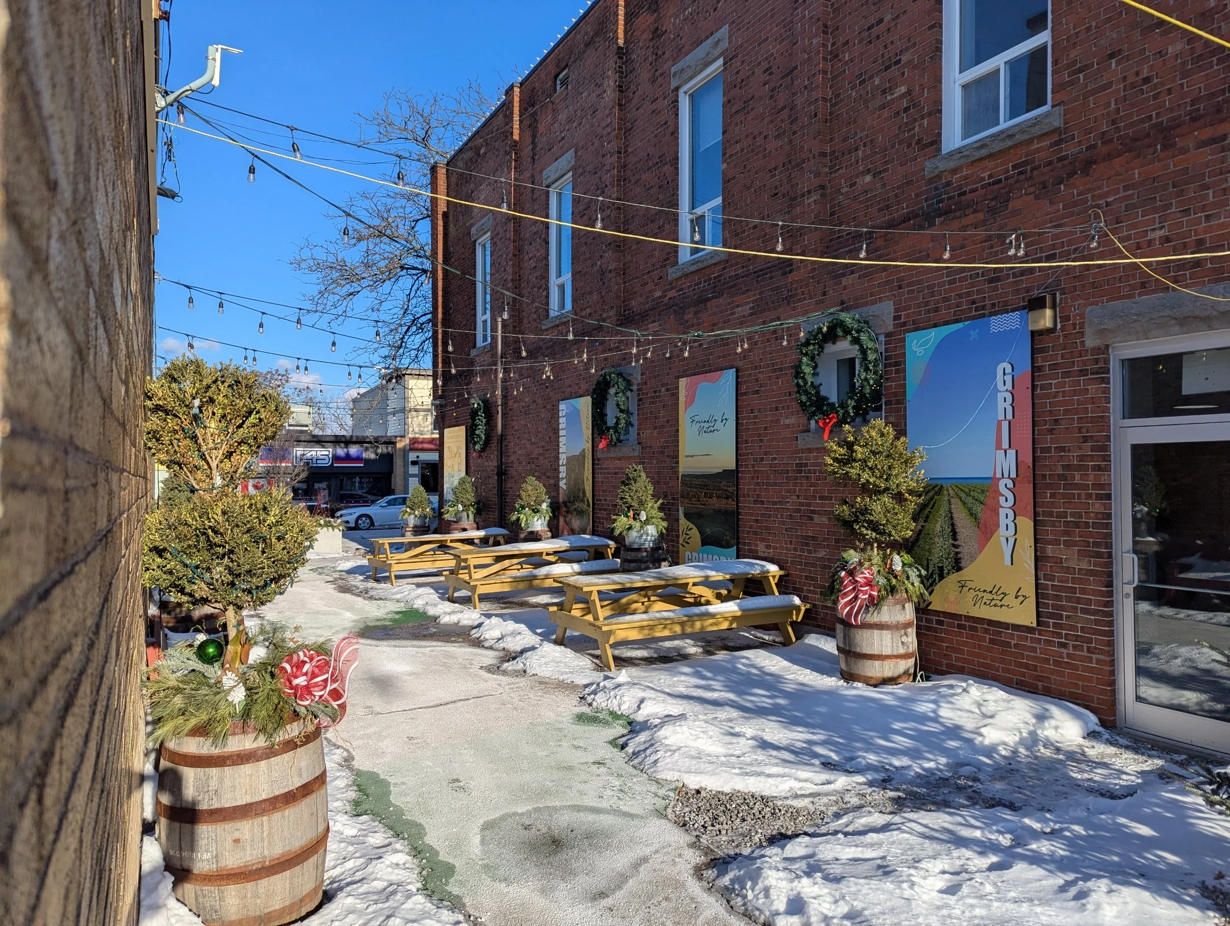 Outdoor patio area decorated for Christmas with wreaths on barrels, small Christmas trees, benches, and string lights hanging over the space outside a brick building with large windows.