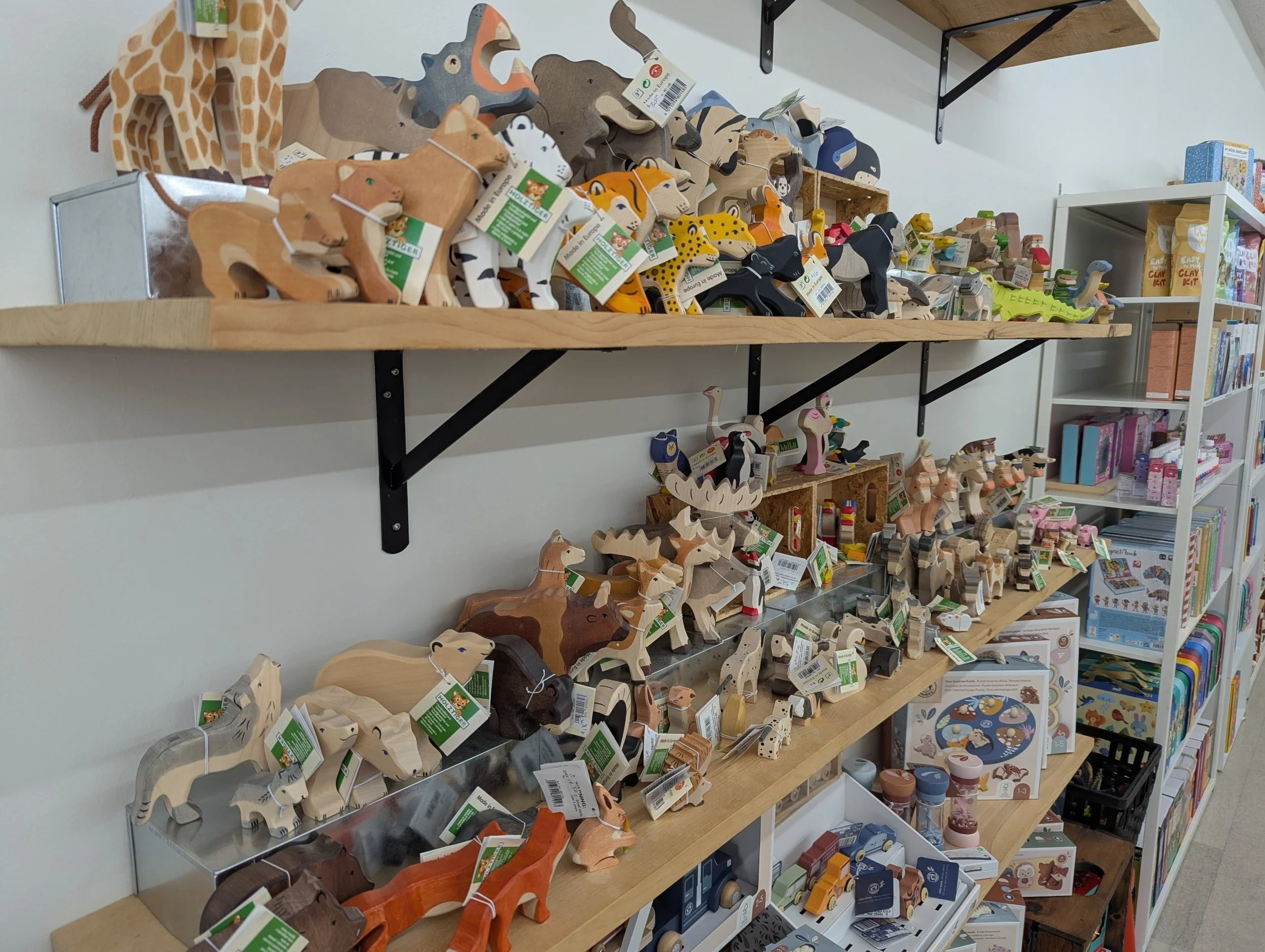 Display shelves stocked with wooden animal toys including giraffes, lions, tigers, elephants, and other animals, with some toys featuring colorful patterns and others in natural wood tones, alongside books and craft kits on nearby shelves.