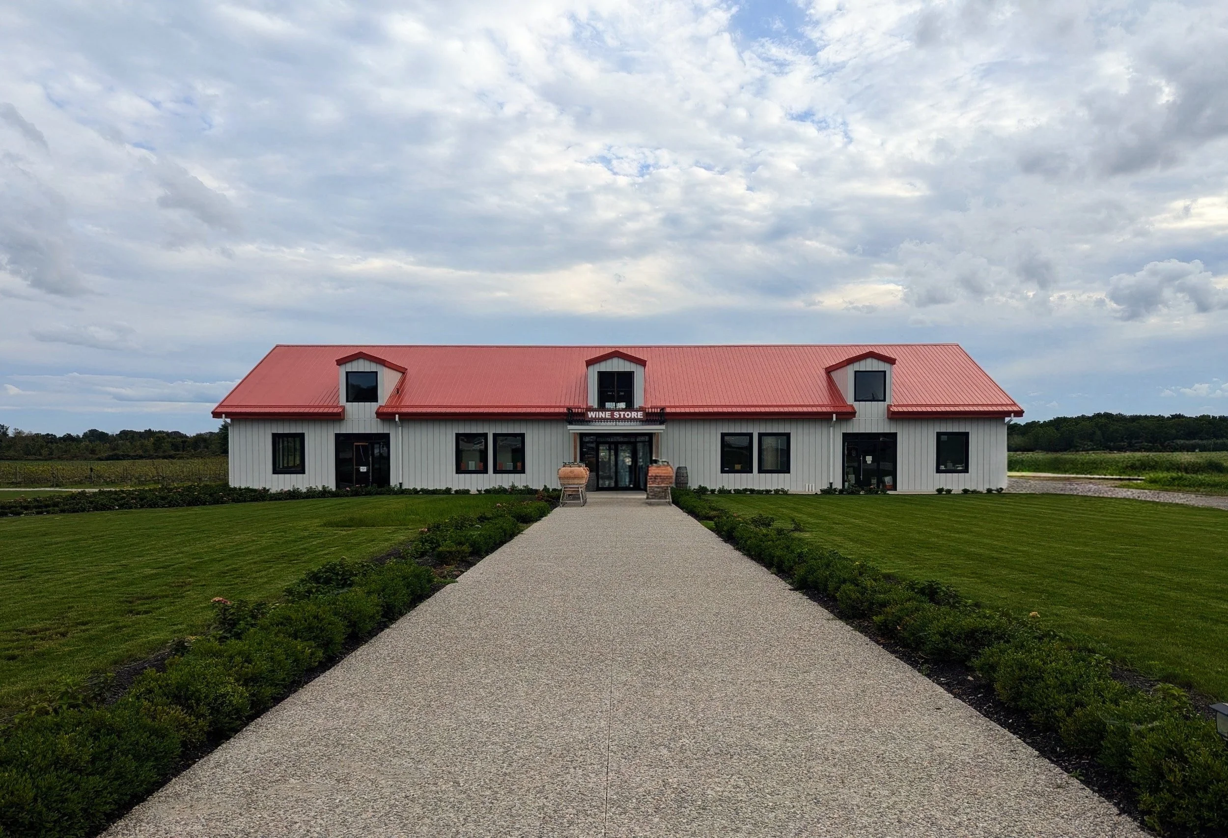 White building with a red roof labeled 'Wine Store,' surrounded by green lawns and a gravel pathway leading to the entrance, with cloudy sky above.