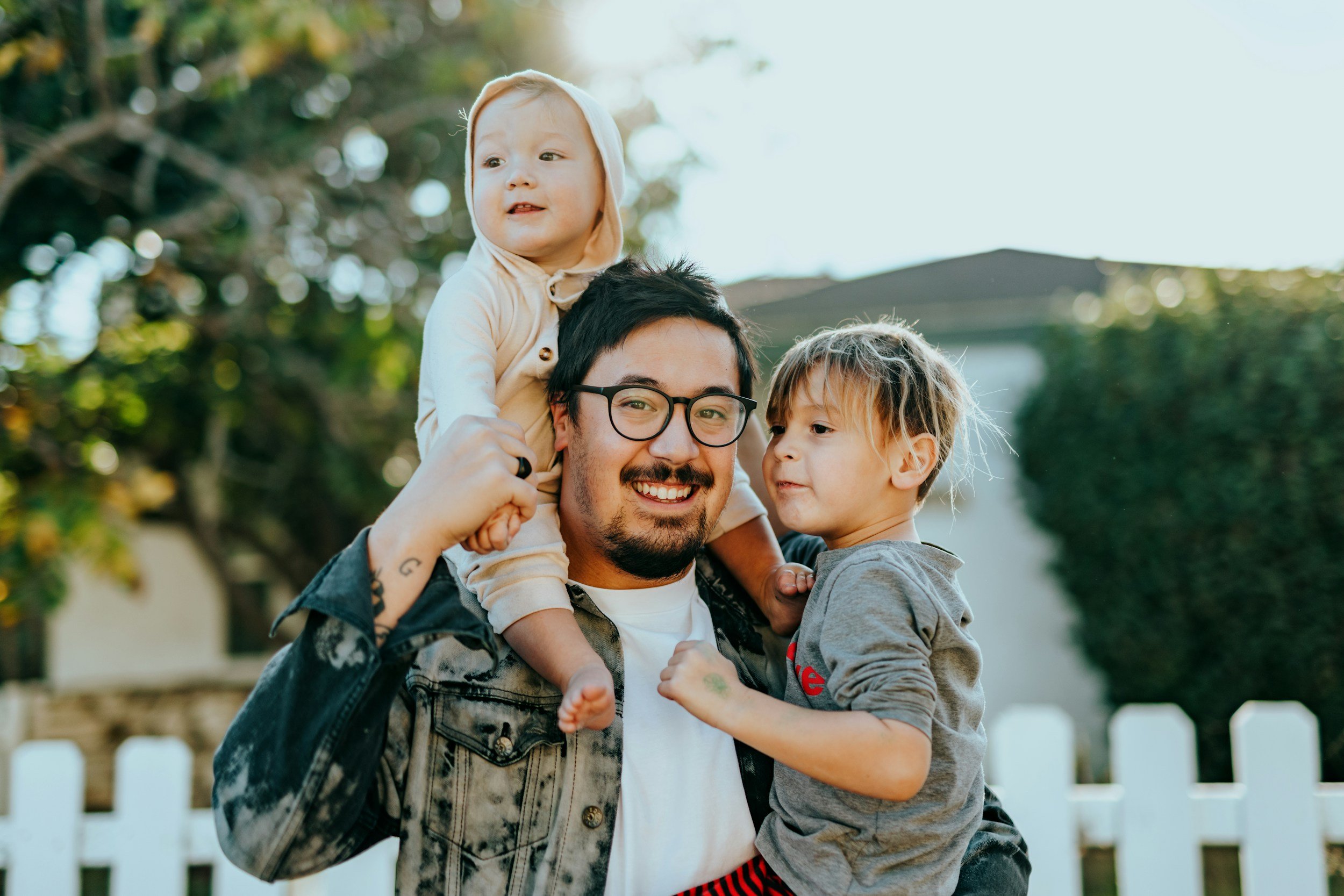 A smiling man with glasses and tattoos carrying a young girl on his shoulders, while a boy leans on his arm, outdoors in front of a white picket fence and trees.