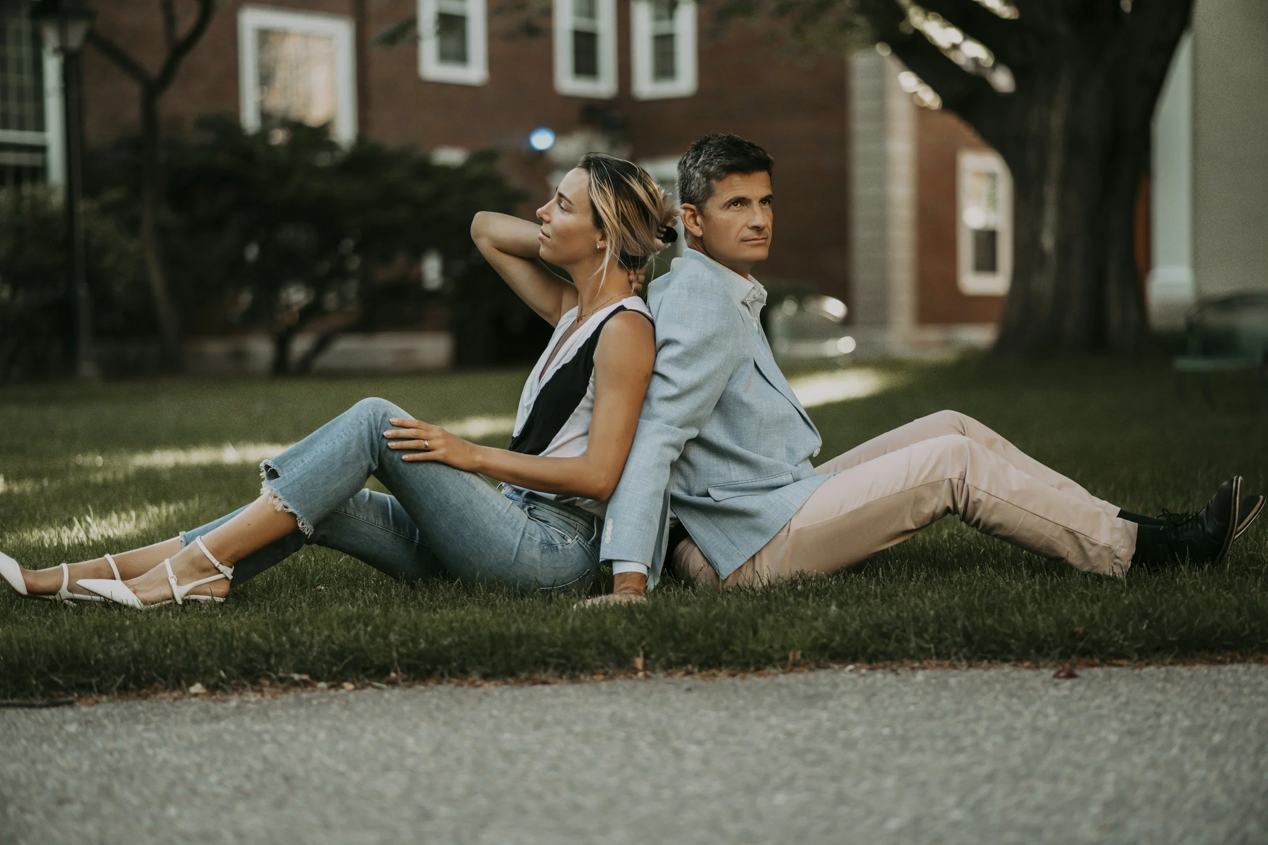 A man and woman sitting back-to-back on the grass in a park, with a house and trees in the background.