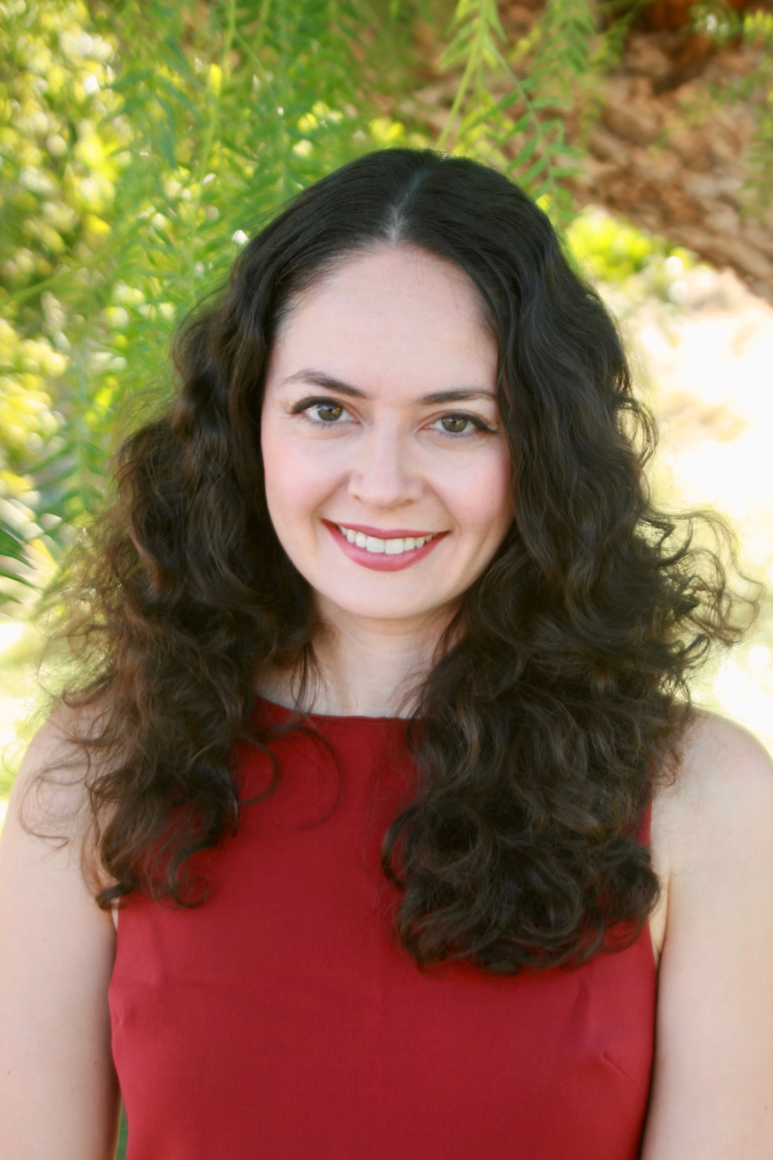 A woman with long, dark, wavy hair smiling, wearing a sleeveless red top, outdoors with green foliage in the background.