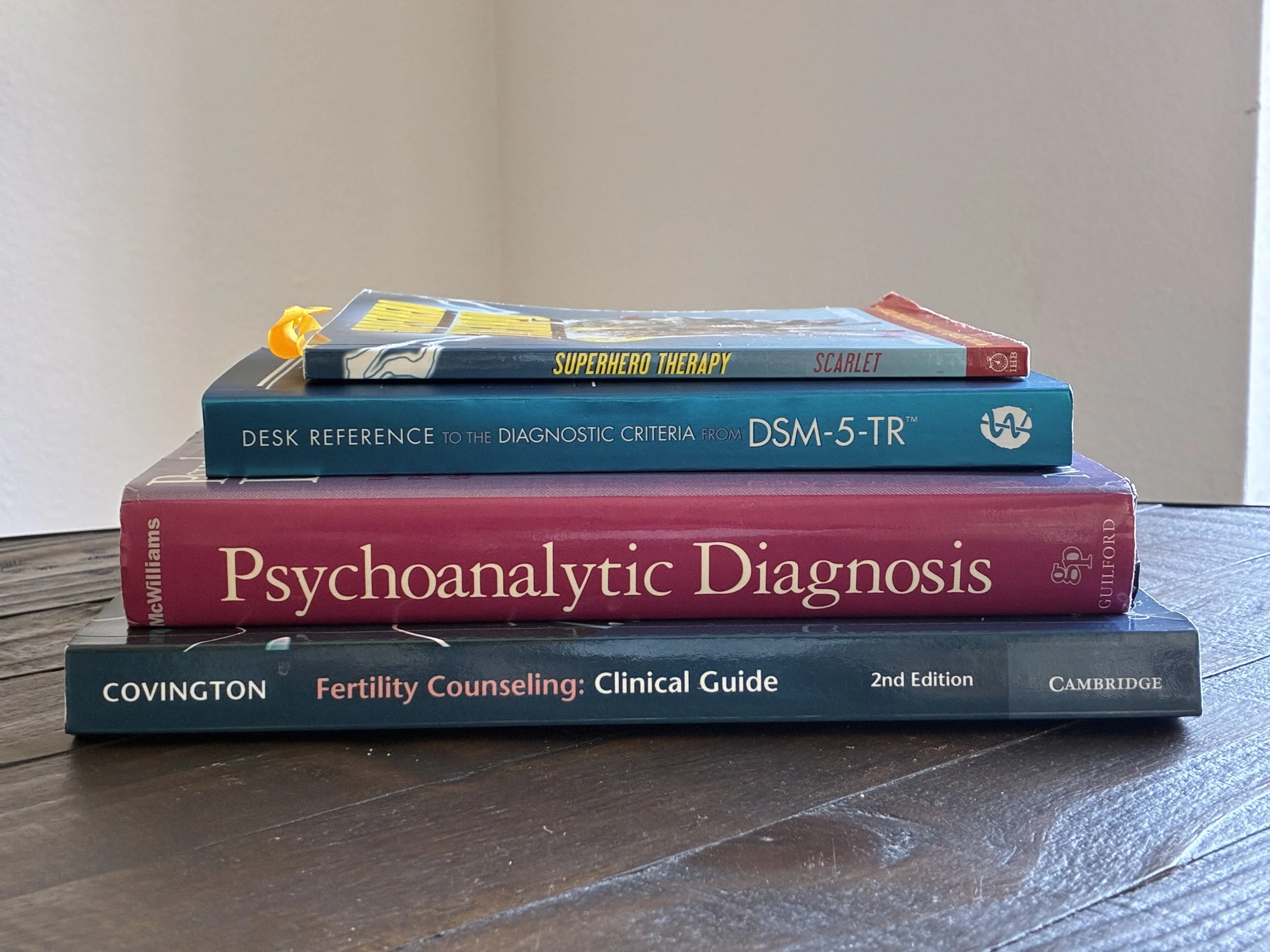 Stack of four books related to psychology and counseling, arranged on a wooden surface. Titles include 'Superhero Therapy', 'DSM-5-TR', 'Psychoanalytic Diagnosis', and 'Fertility Counseling: Clinical Guide'.