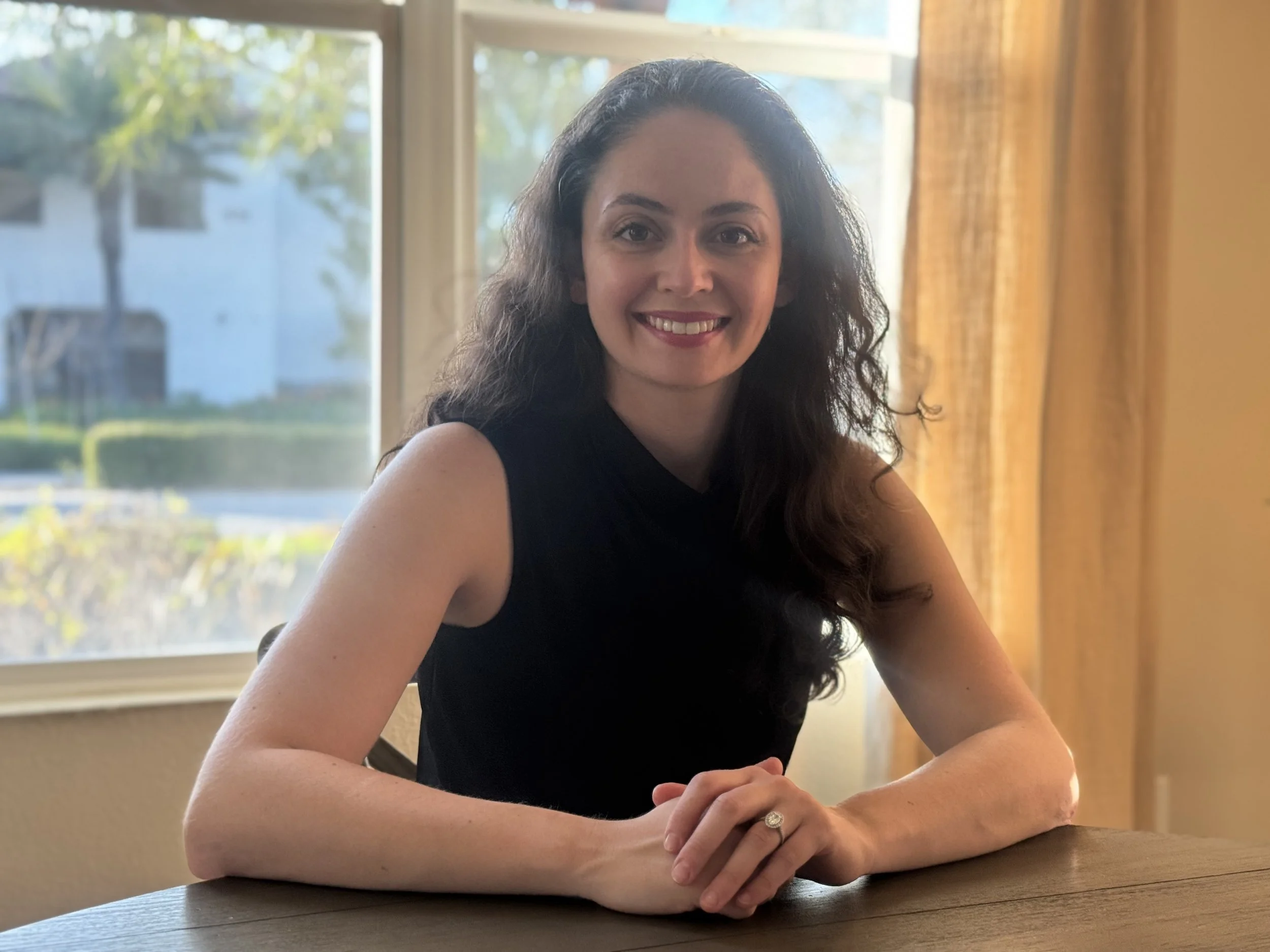 A woman with long, curly dark hair sitting at a wooden table in front of a window, smiling at the camera. She is wearing a sleeveless black top and a ring on her left hand. Bright natural light fills the room.