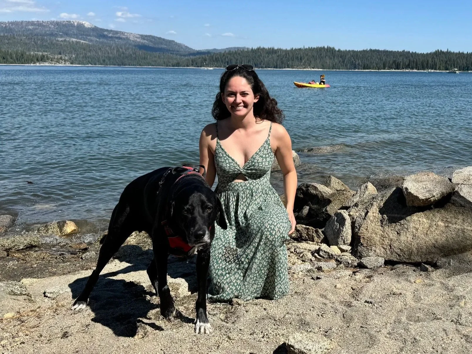 A woman in a green patterned dress kneeling on a sandy beach next to a black dog. The woman is smiling at the camera. Background shows a lake with a person kayaking, a forested shoreline, and mountains under a clear blue sky.
