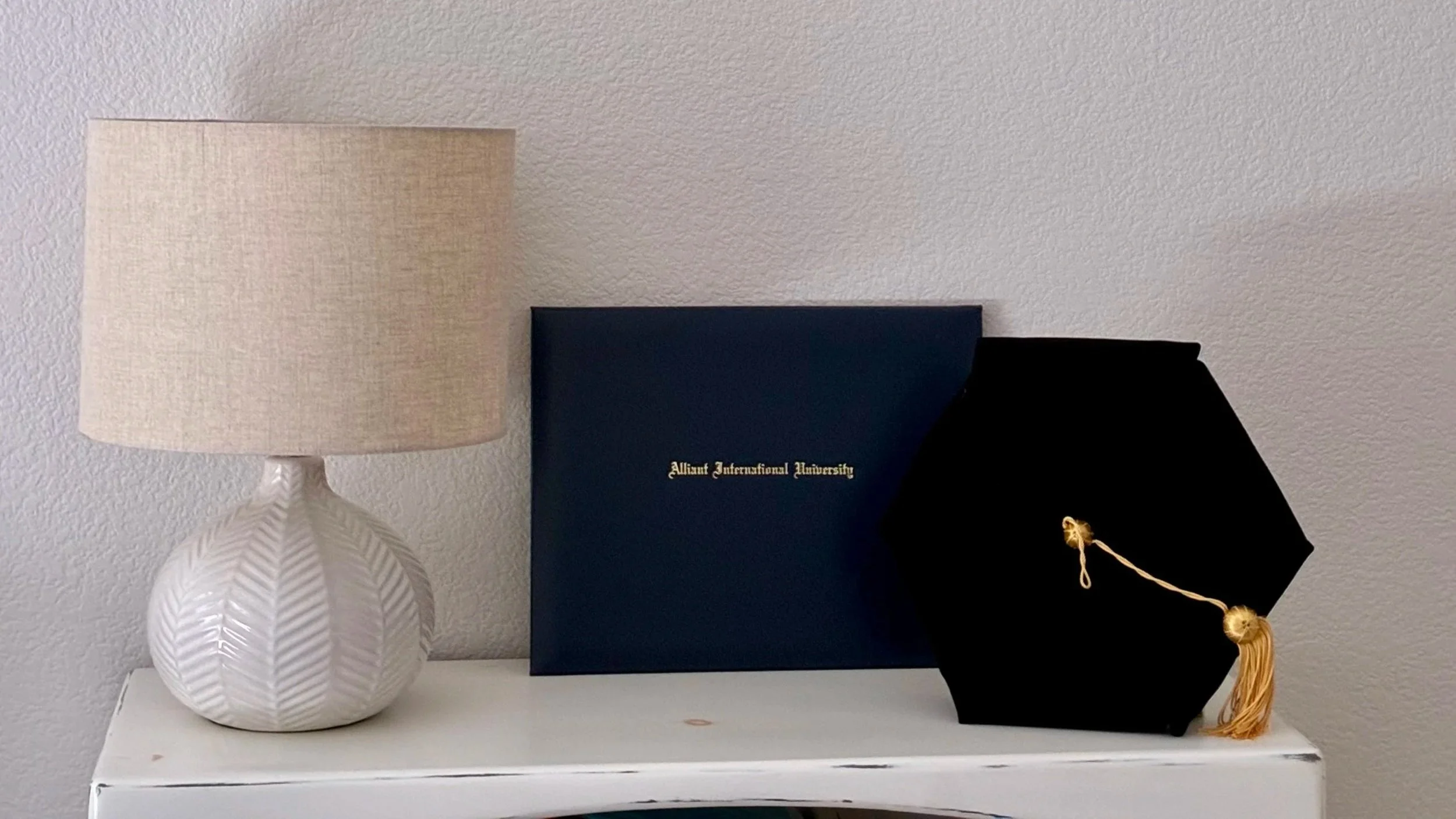 A table with a white ceramic lamp, a blue diploma cover with gold writing, and a black graduation cap with a tassel.