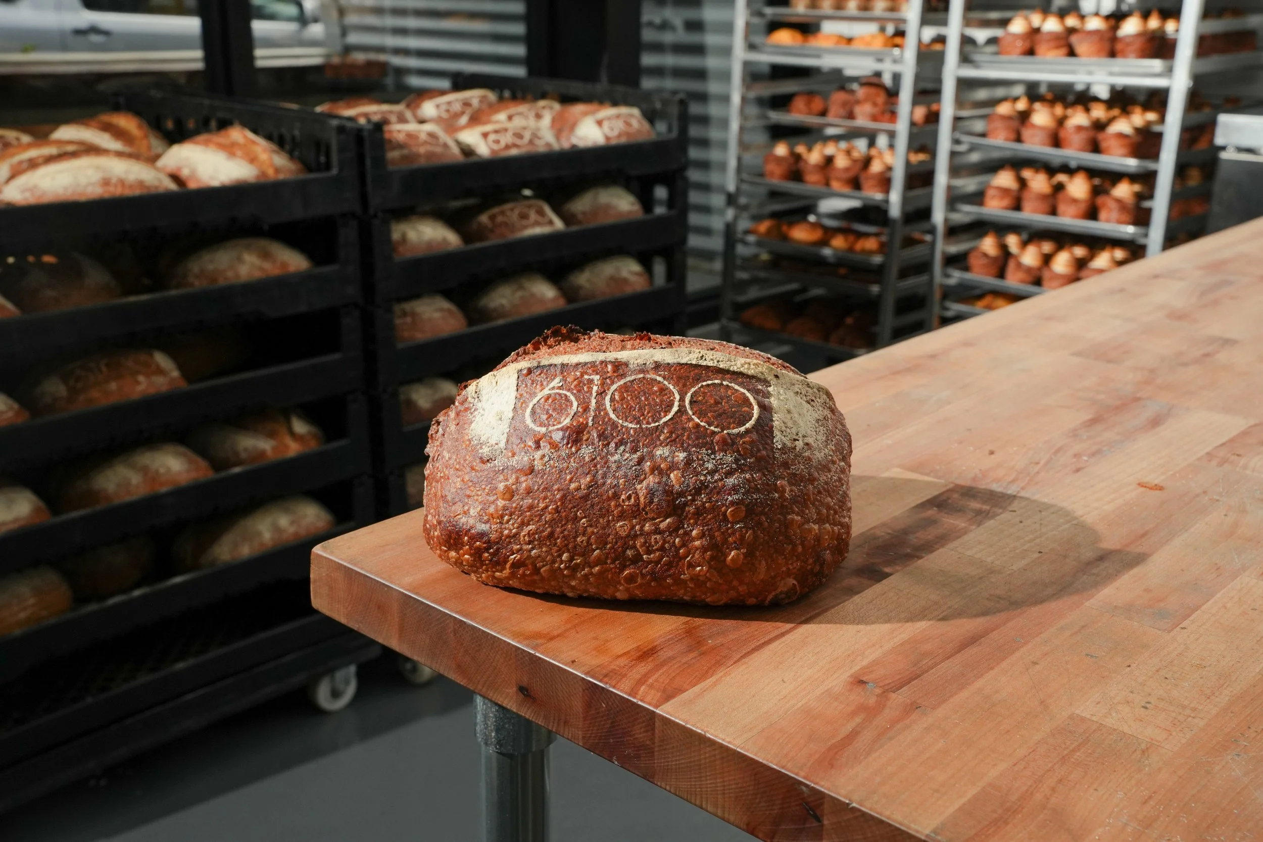 A loaf of bread with the number '6100' baked into the crust, placed on a wooden table in a bakery or bread-making facility with racks of baked bread in the background.