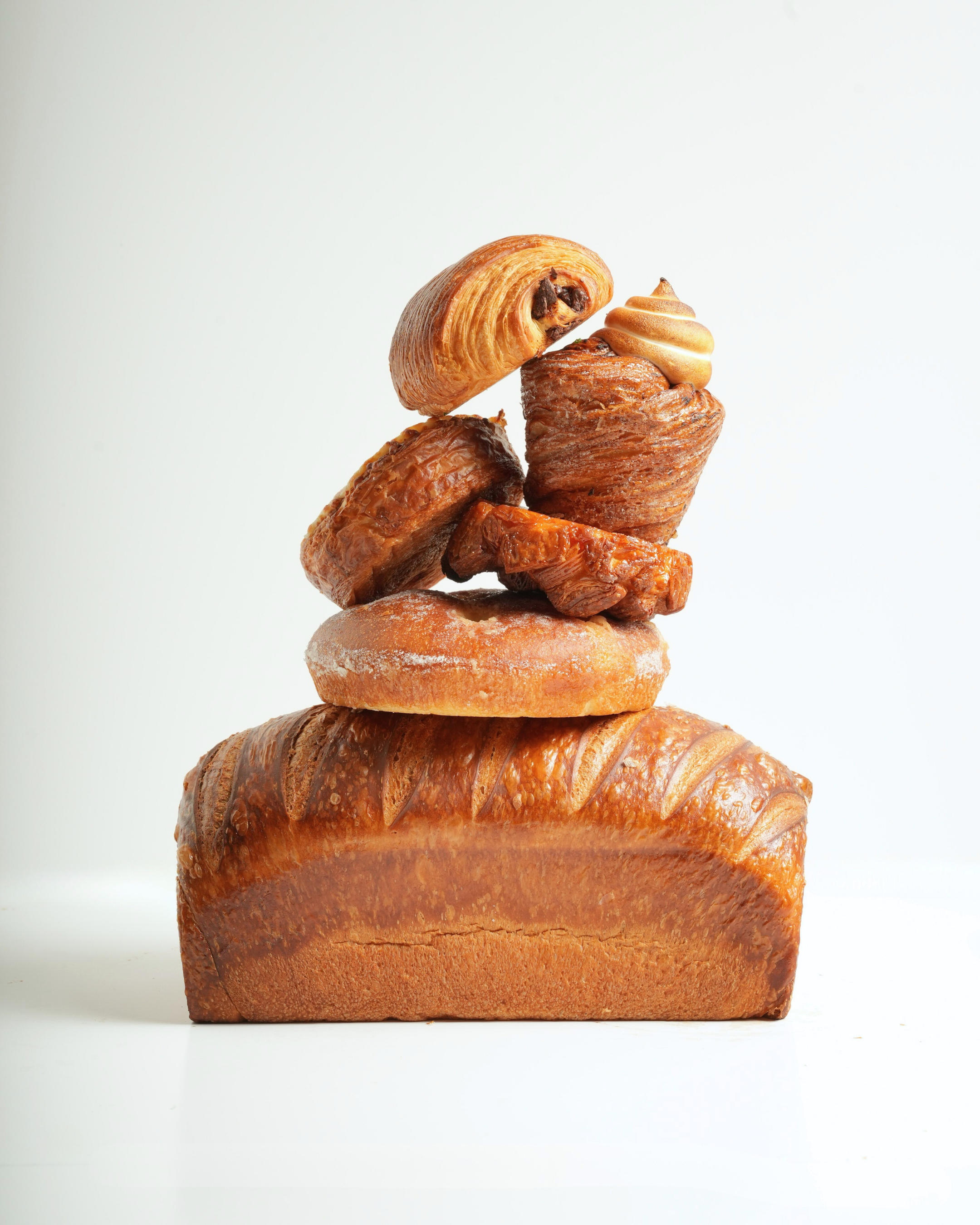 Stacked assorted baked goods including bread, croissant, pastry, and cupcakes against a white background.