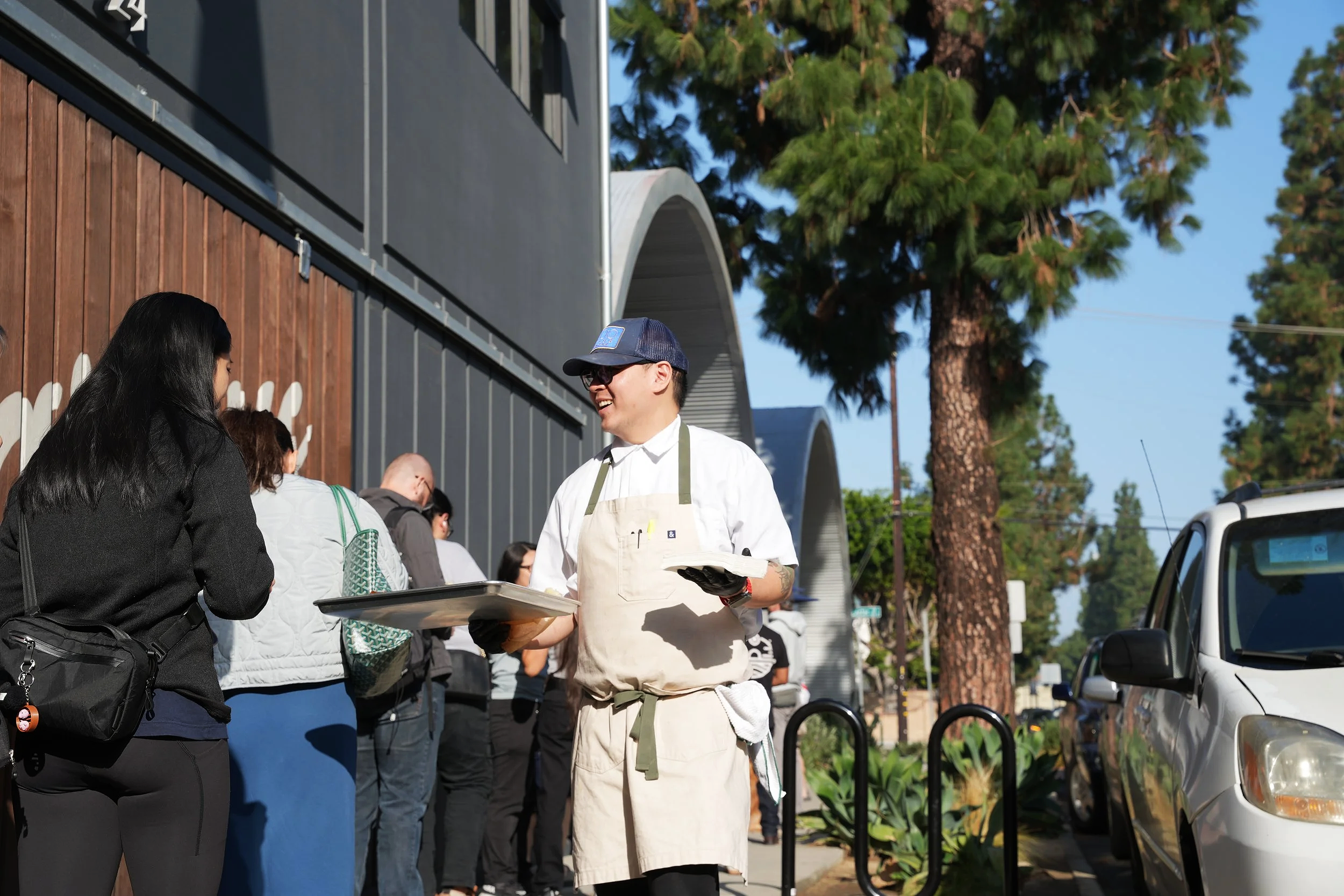 A smiling man in a white apron and cap holding a tray and a menu talking to a woman with a black jacket outside a restaurant or cafe.