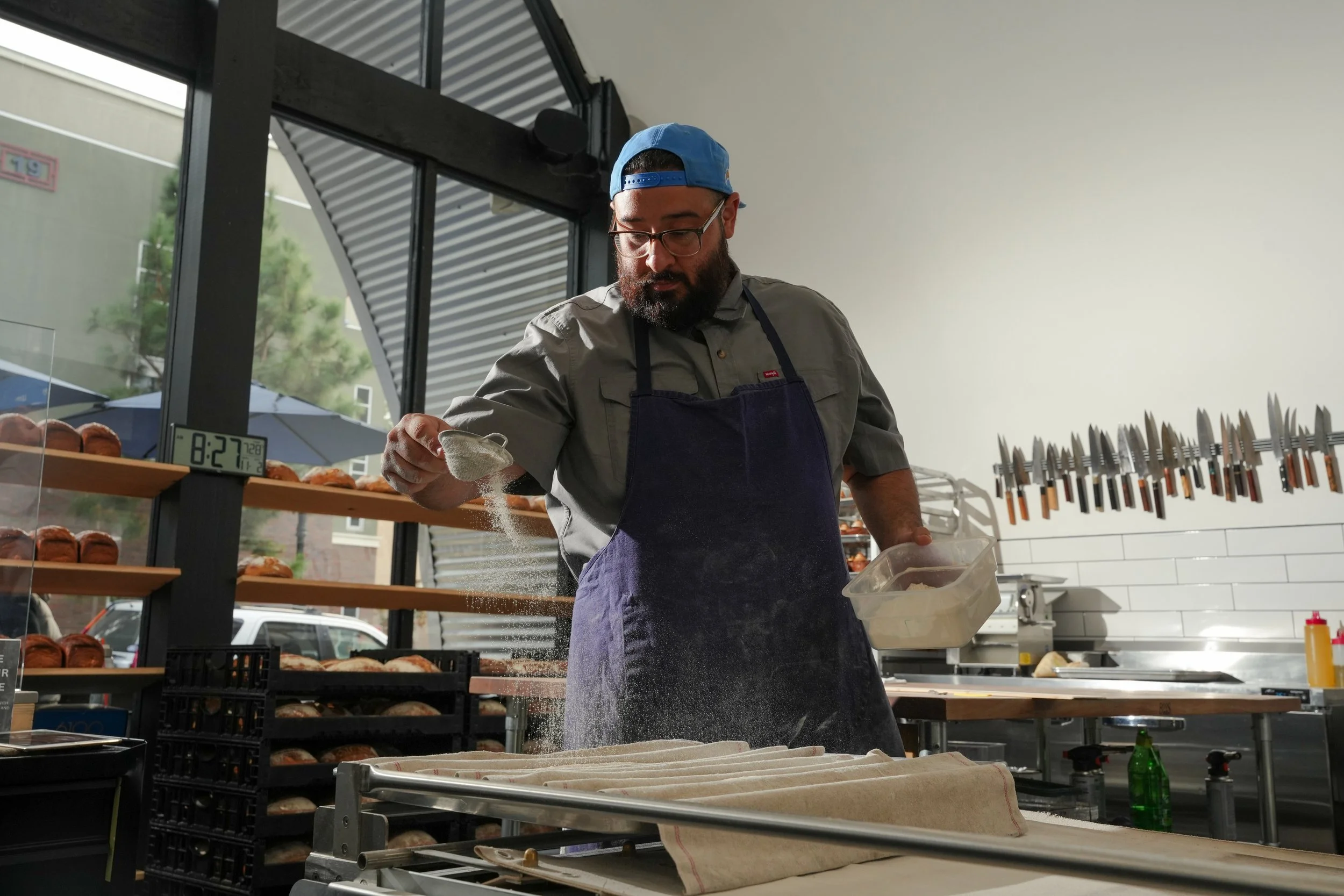 A man with glasses, a beard, wearing a blue cap, gray shirt, and black apron sprinkling flour onto a dough in a bakery or kitchen.