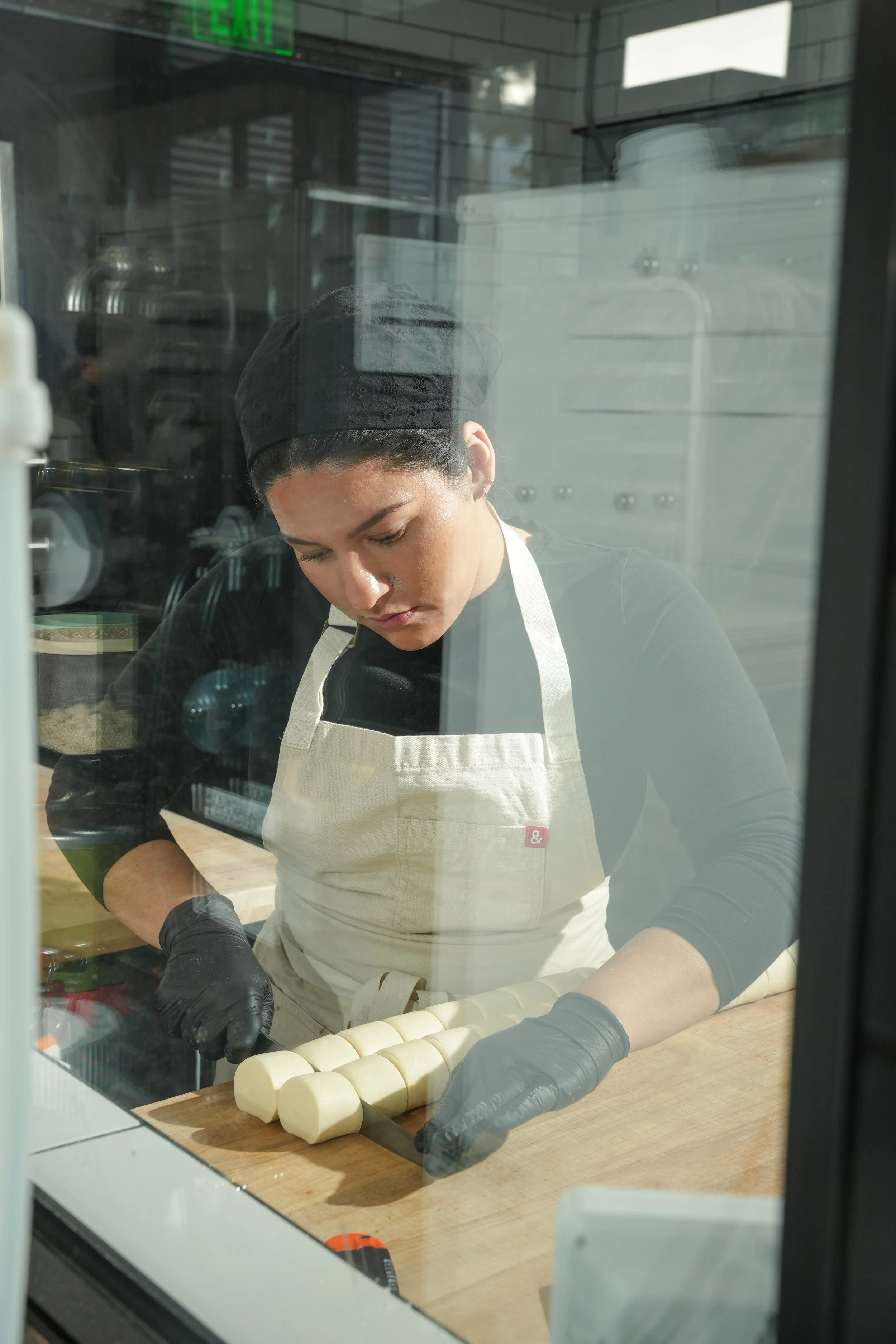 A woman wearing a black cap, black gloves, and a cream apron is slicing a rolled piece of dough on a wooden surface inside a commercial kitchen, viewed through a glass window.