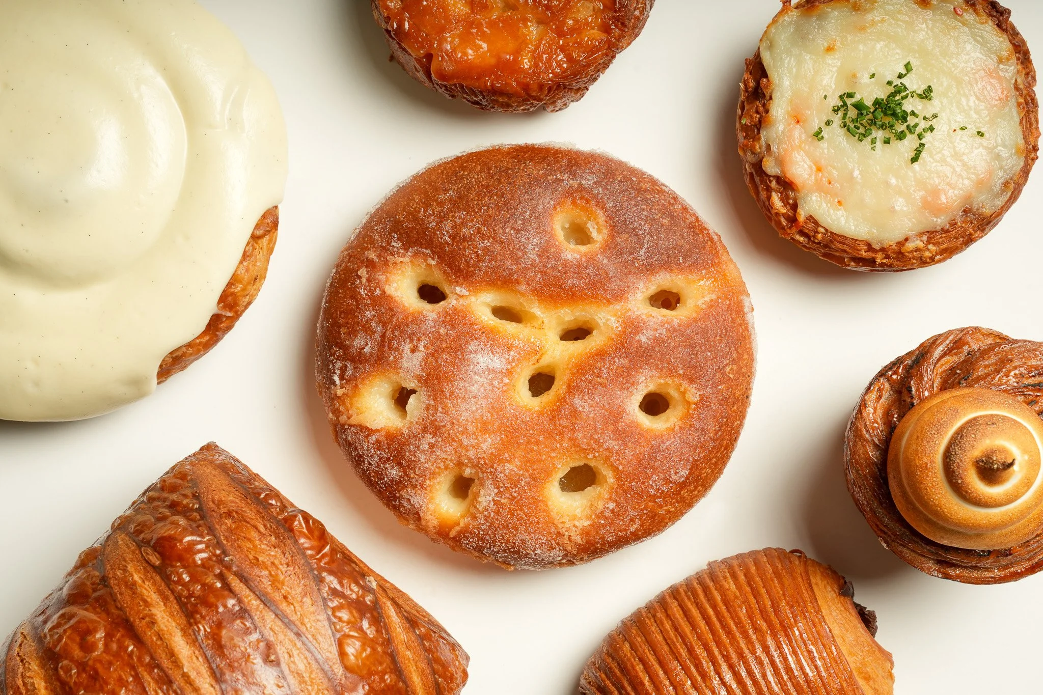 Assorted baked goods including a round bread roll with holes, a croissant, a cinnamon roll, a Danish pastry, and two cheese or savory muffins on a white surface.