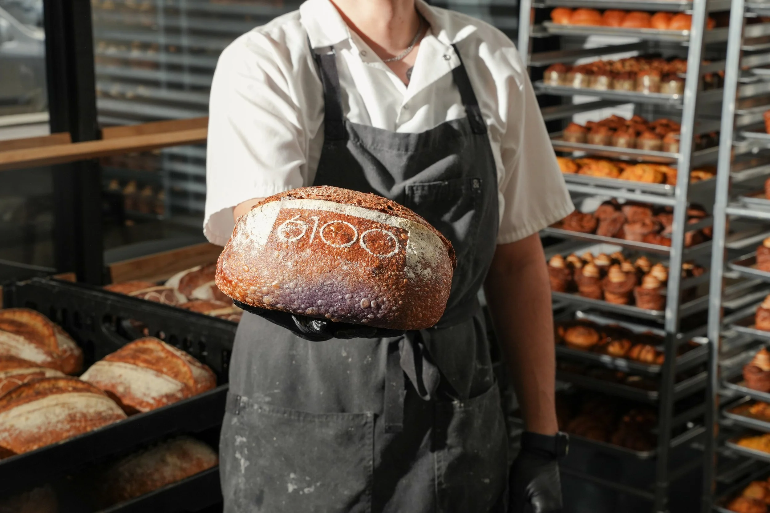 Person in white shirt and black apron holding a loaf of bread with the word 'bloom' stamped on it, in a bakery with shelves of baked goods in the background.