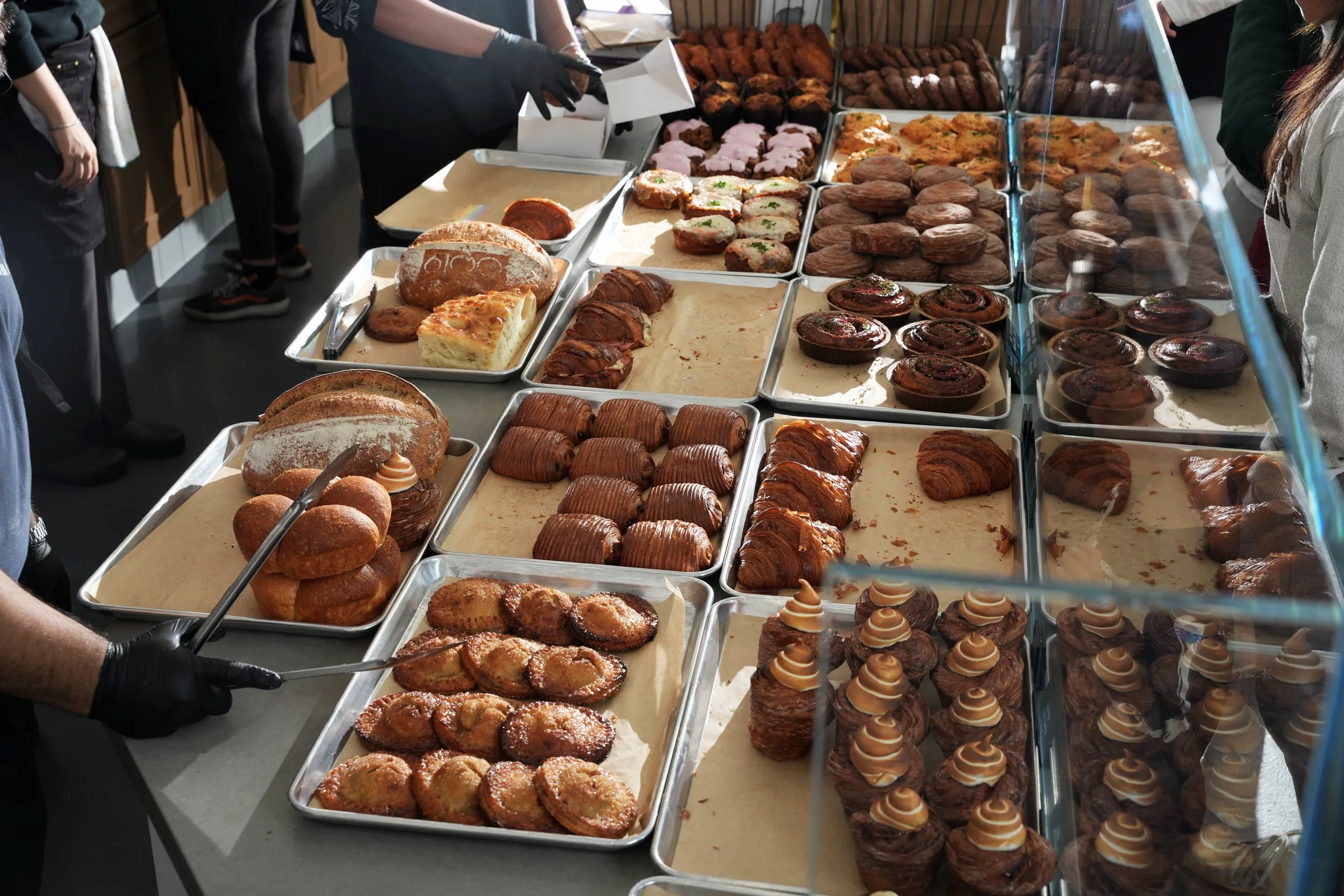 A bakery display with trays of various pastries, including croissants, muffins, danishes, and other baked goods, with some people selecting treats.