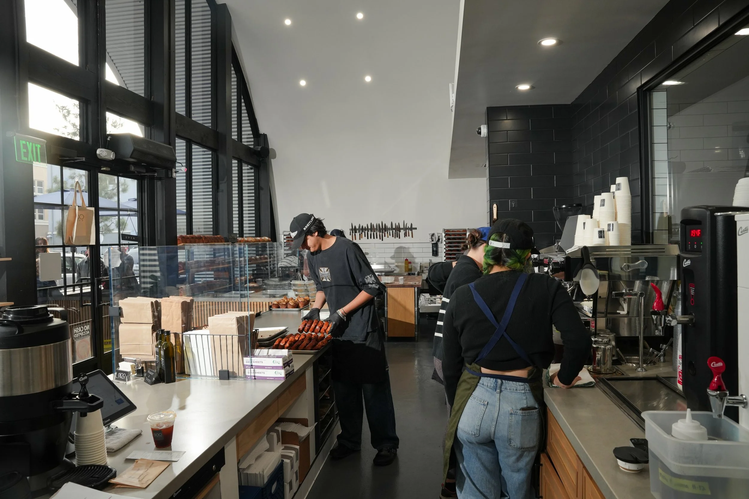 Inside a modern cafe, baristas prepare coffee and food behind the counter, with large windows letting in natural light and a visible street outside.