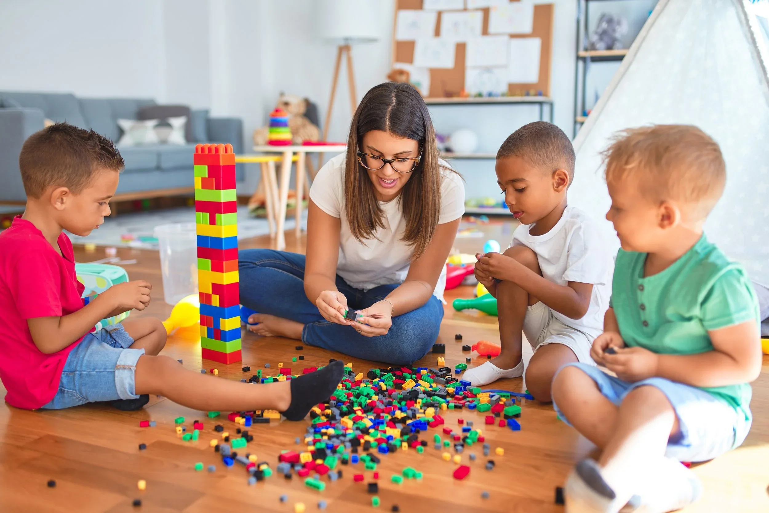 A woman and three young children playing with colorful building blocks on a wooden floor in a playroom.