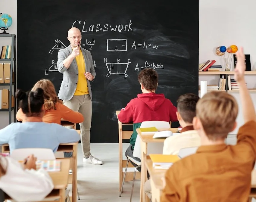 Teacher standing in front of blackboard with math formulas, teaching students in classroom, one student raising hand.