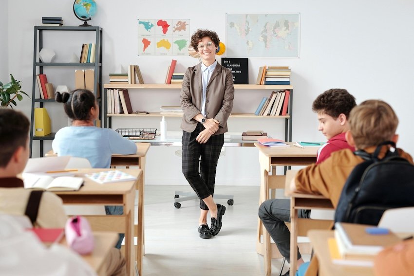 A teacher standing at the front of a classroom with students seated at desks. The teacher is smiling, wearing glasses and a gray blazer. The classroom has bookshelves, globe, and world maps on the wall.