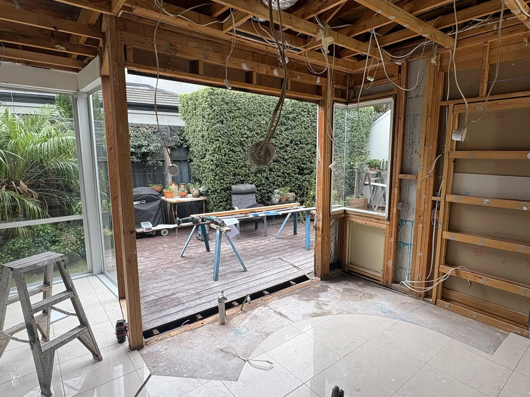 View of a Melbourne house under renovation, with wooden framing and exposed wiring inside, and a sliding glass door leading to an outdoor patio.