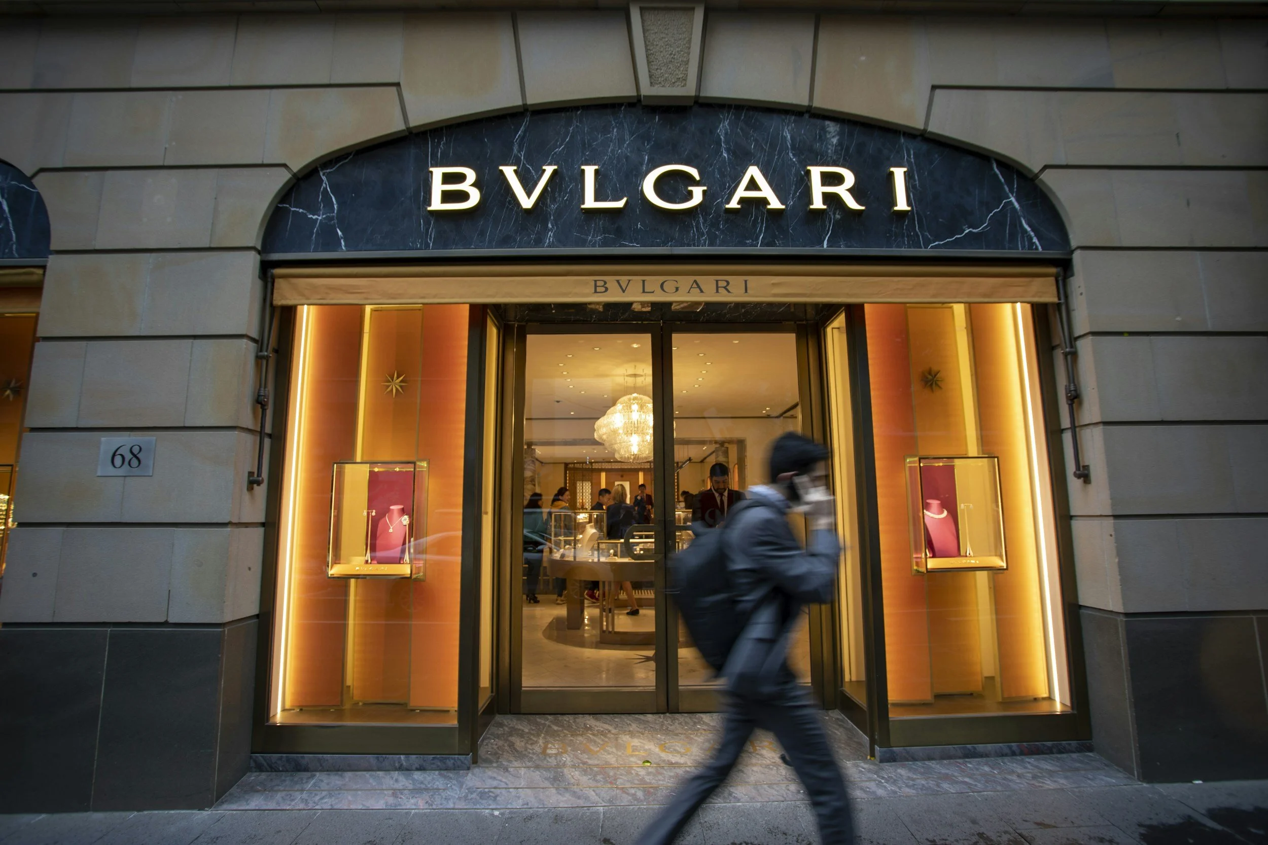 Storefront entrance of Bvlgari jewelry store with illuminated sign, large display windows showing jewelry displays, and a person walking past outside.