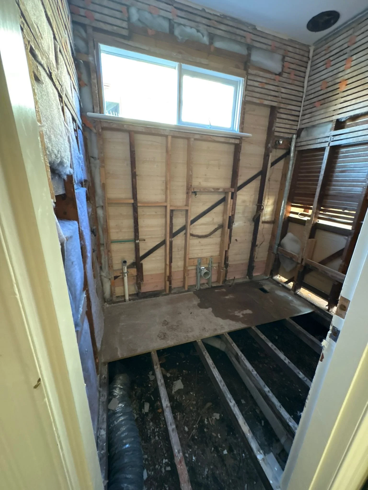 A bathroom under renovation with exposed wooden wall framing, plumbing pipes, a floor with visible subflooring and floor joists, and a window providing natural light.