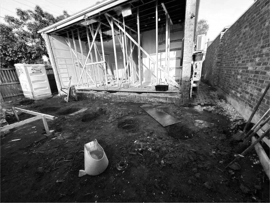 Rear view of a house under renovation, with exposed wooden framing and construction debris in the yard.