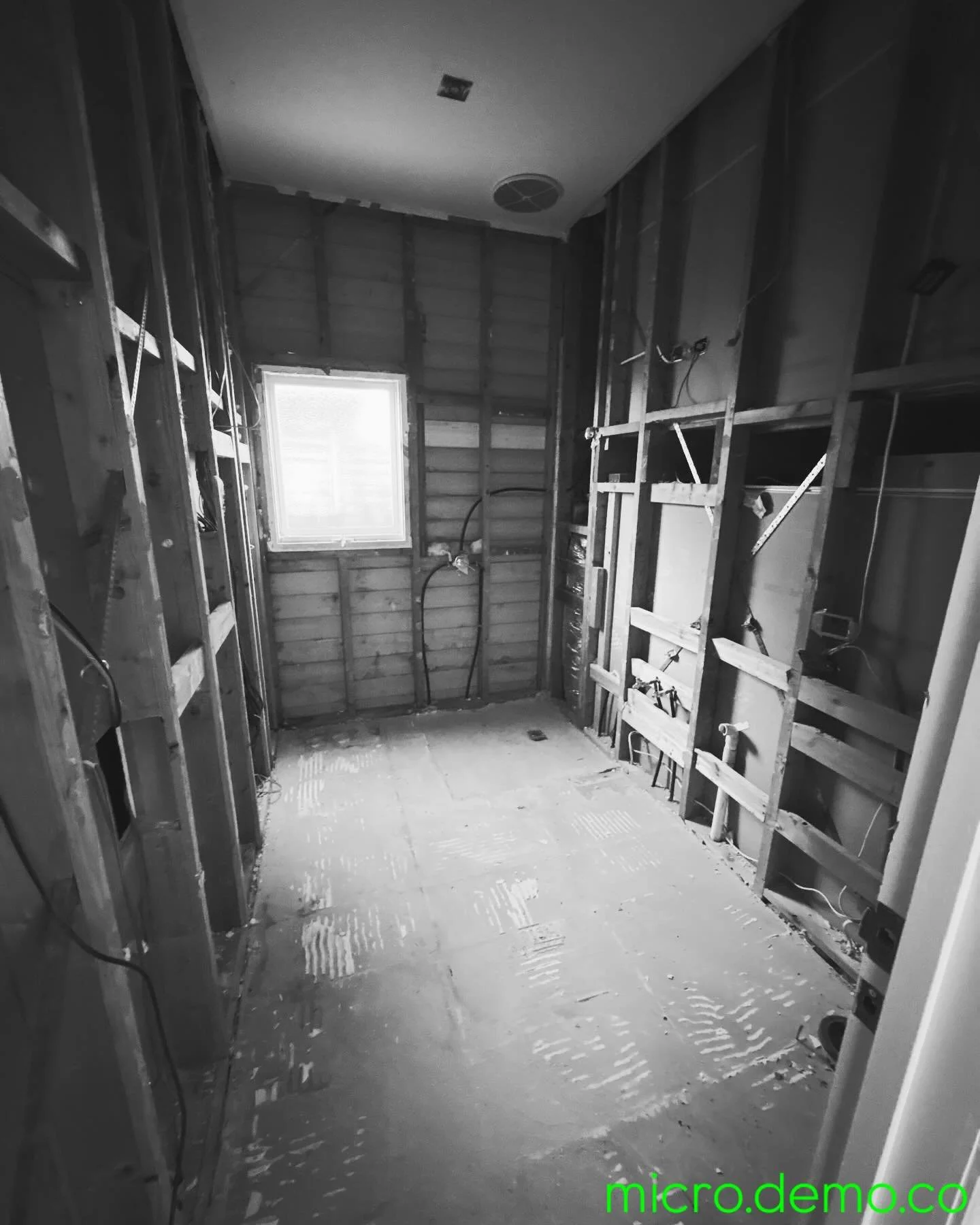 Empty, unfinished room under construction with exposed wooden framing and electrical wiring, small window on the back wall, and a textured concrete floor.