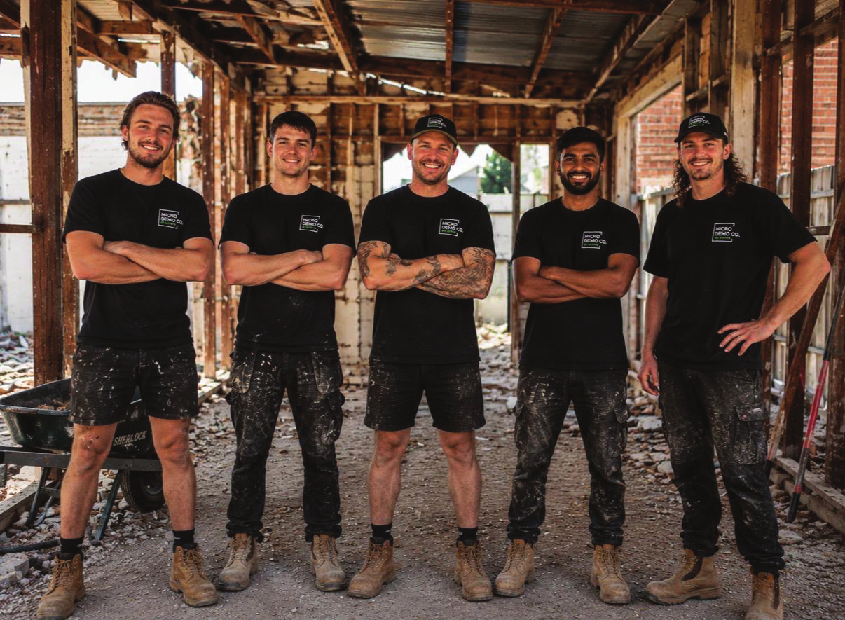 Five construction workers standing with arms crossed inside a partially built structure, smiling at the camera. They are wearing black t-shirts, work pants, and boots, with some having paint splatters on their pants and a tattoo on the middle worker.