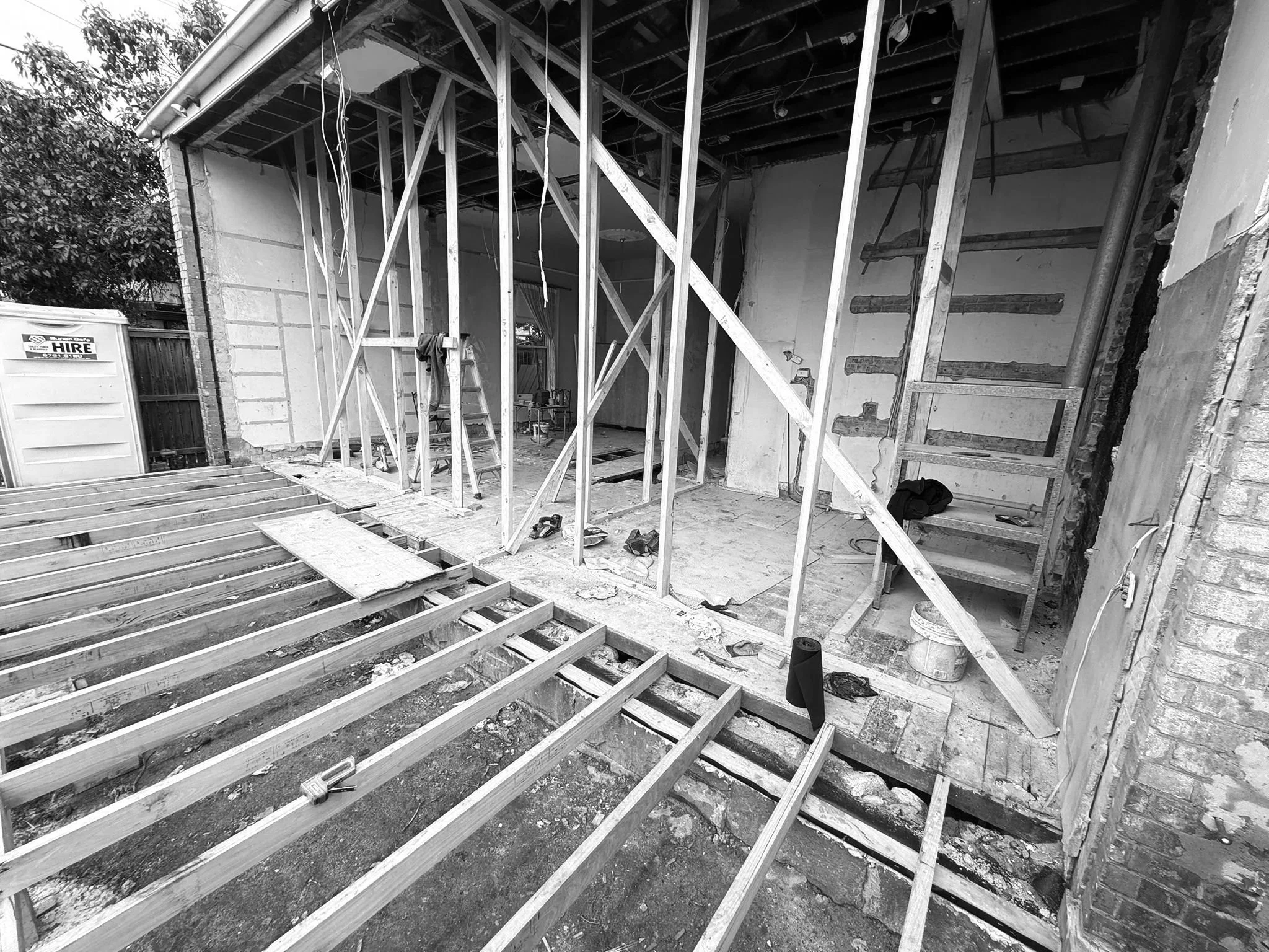 Interior view of a house under construction with wooden framing, a partially built staircase, construction tools, and a portable toilet outside.