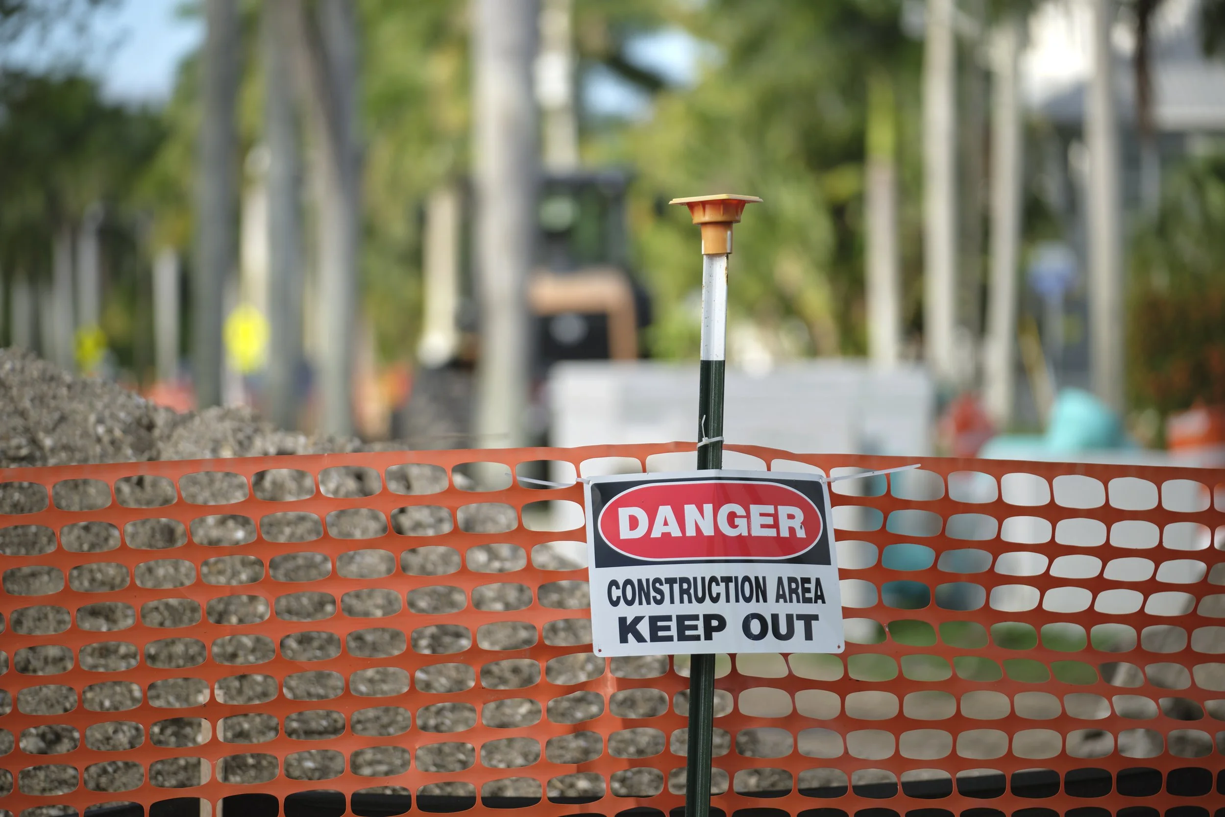 Construction site with orange safety barrier and a warning sign that reads 'Danger Construction Area Keep Out.' The background shows trees and blurry construction equipment.