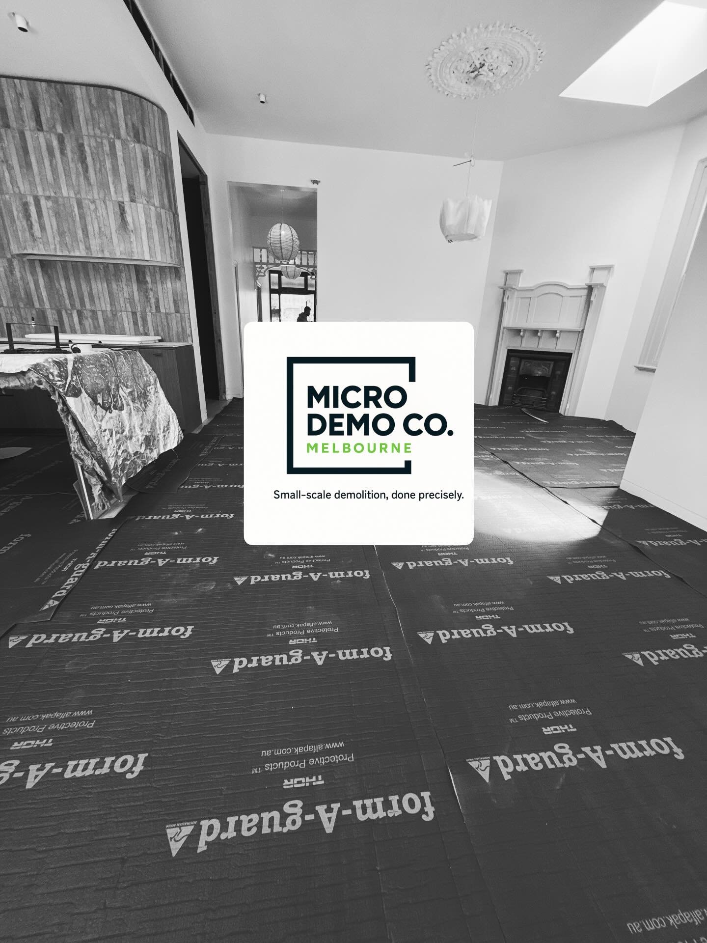 Interior room with flooring marked for demolition, a covered table, and a sign reading "Micro Demol Co. Melbourne - Small-scale demolition, done precisely." a doorway leads to another room, with a person visible in the background.