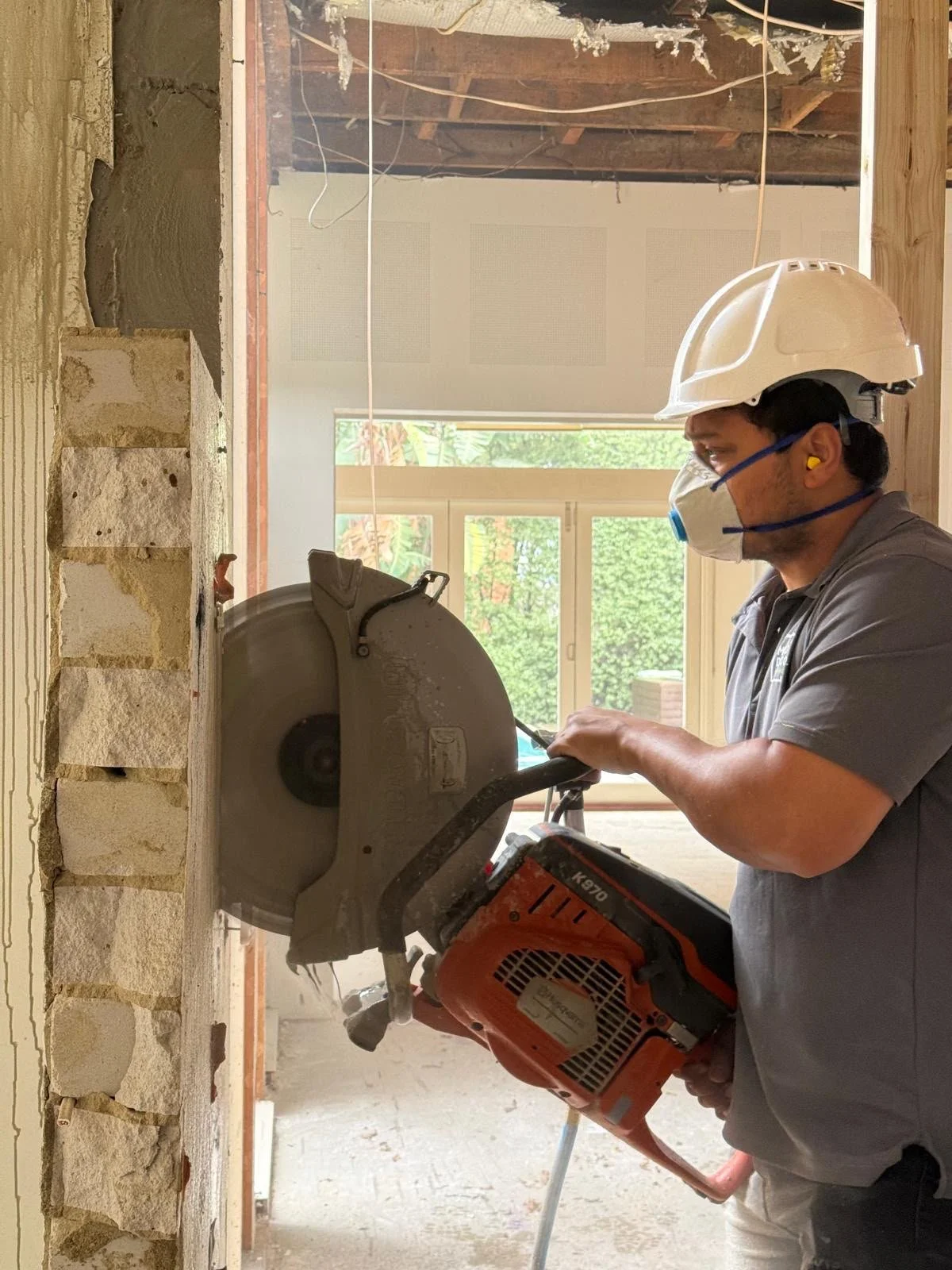A construction worker is using a large chop saw to cut a block of stone or concrete. He is wearing a white safety helmet, a gray face mask, and yellow ear protection. The room has unfinished walls and a window in the background with a view of trees outside.