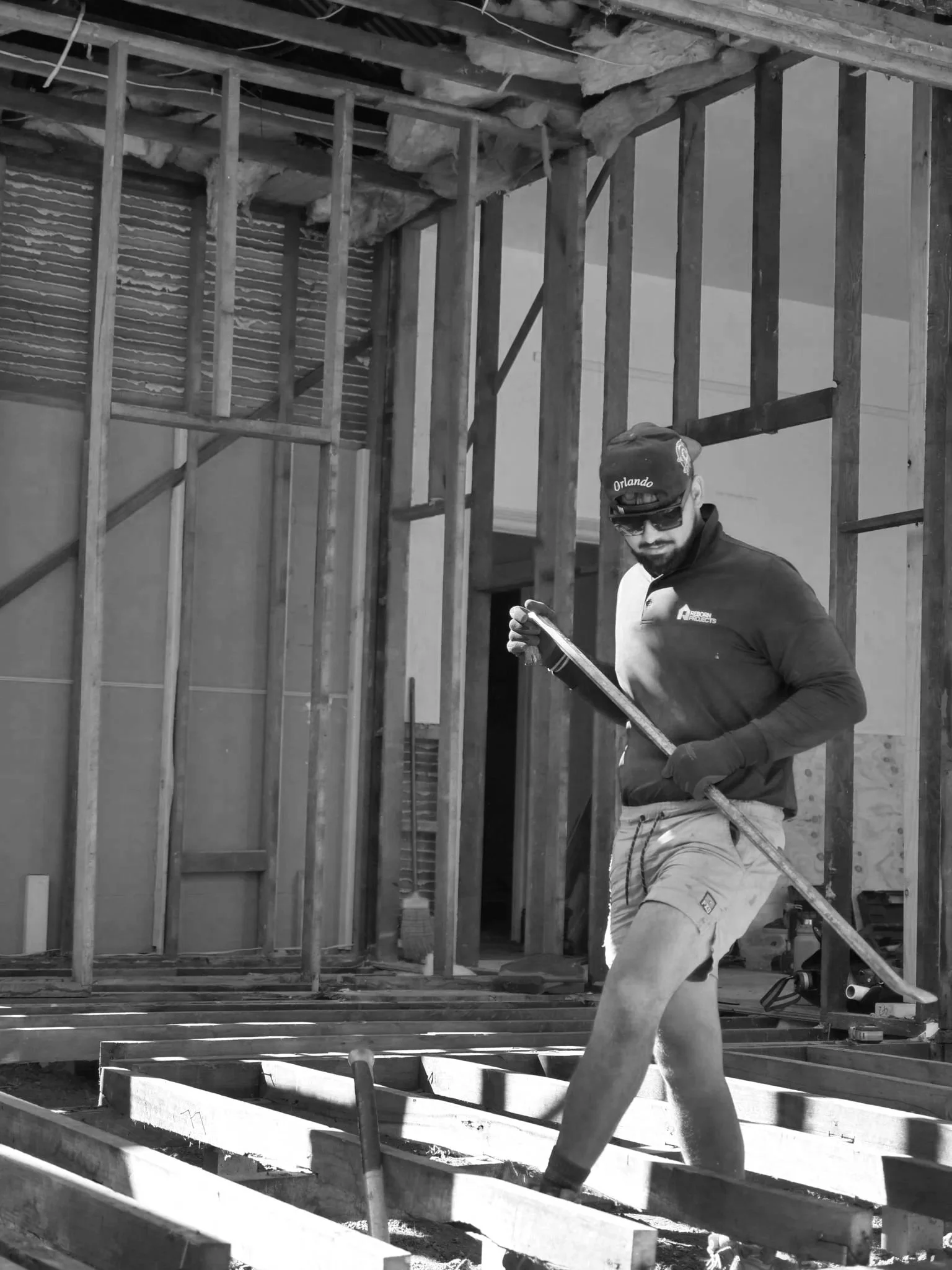 A man wearing sunglasses, a cap with 'Orlando' on it, a jacket, gloves, and shorts working with a tool on a construction site with exposed wooden framing.
