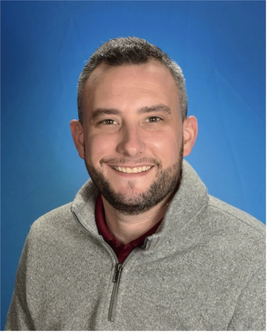 A smiling young man with short brown hair and a beard, wearing a light gray zip-up sweater and a maroon shirt underneath, posed against a blue background.