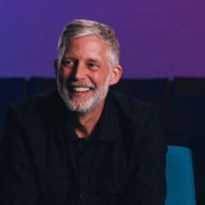 A smiling man with gray hair and a beard, wearing a black shirt, sitting against a dark background with purple lighting.