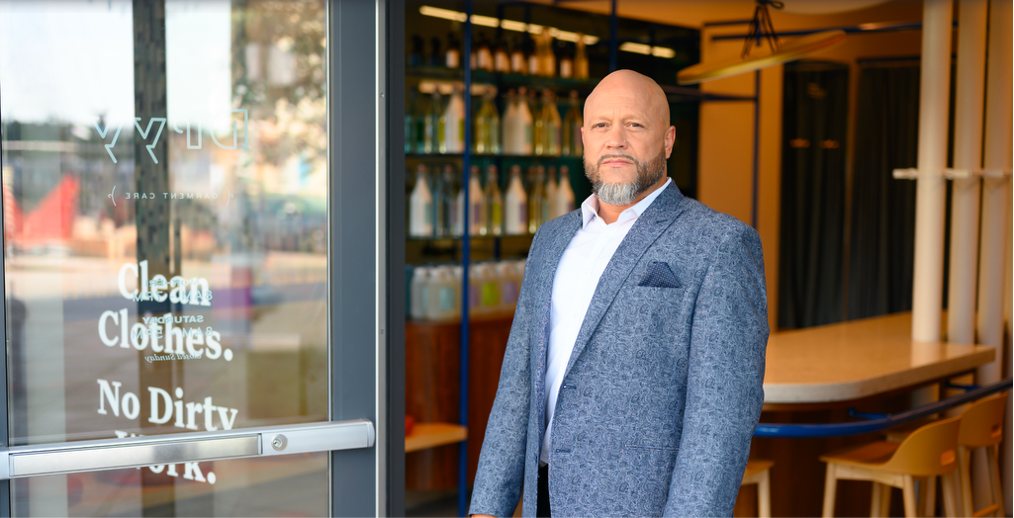 A man in a blue patterned blazer and white shirt standing outside a store with glass doors, with shelves of bottles inside.