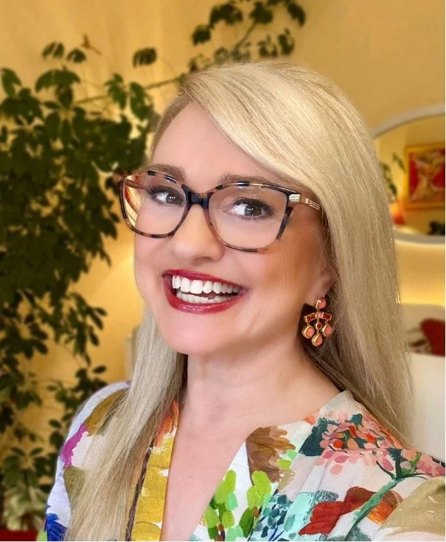 A woman with long blonde hair, wearing glasses, colorful floral top, and pink earrings, smiling indoors with a leafy plant in the background.