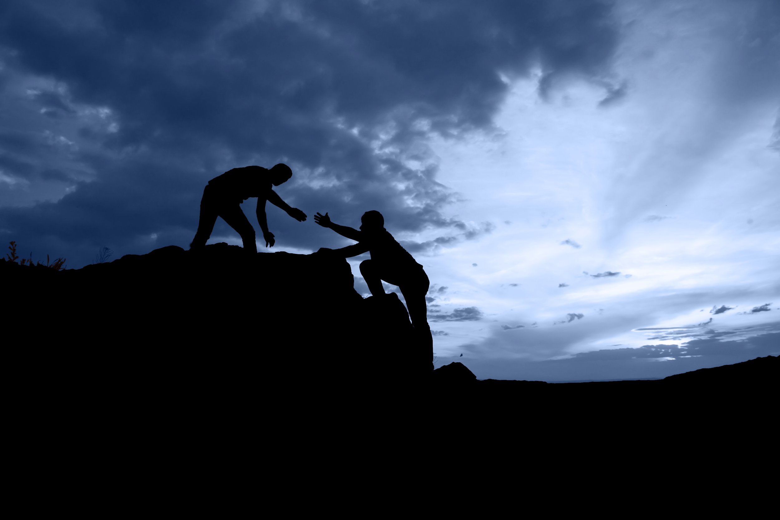 Two people silhouetted against a cloudy sky on a rocky hilltop; one is helping the other climb up by extending a hand.