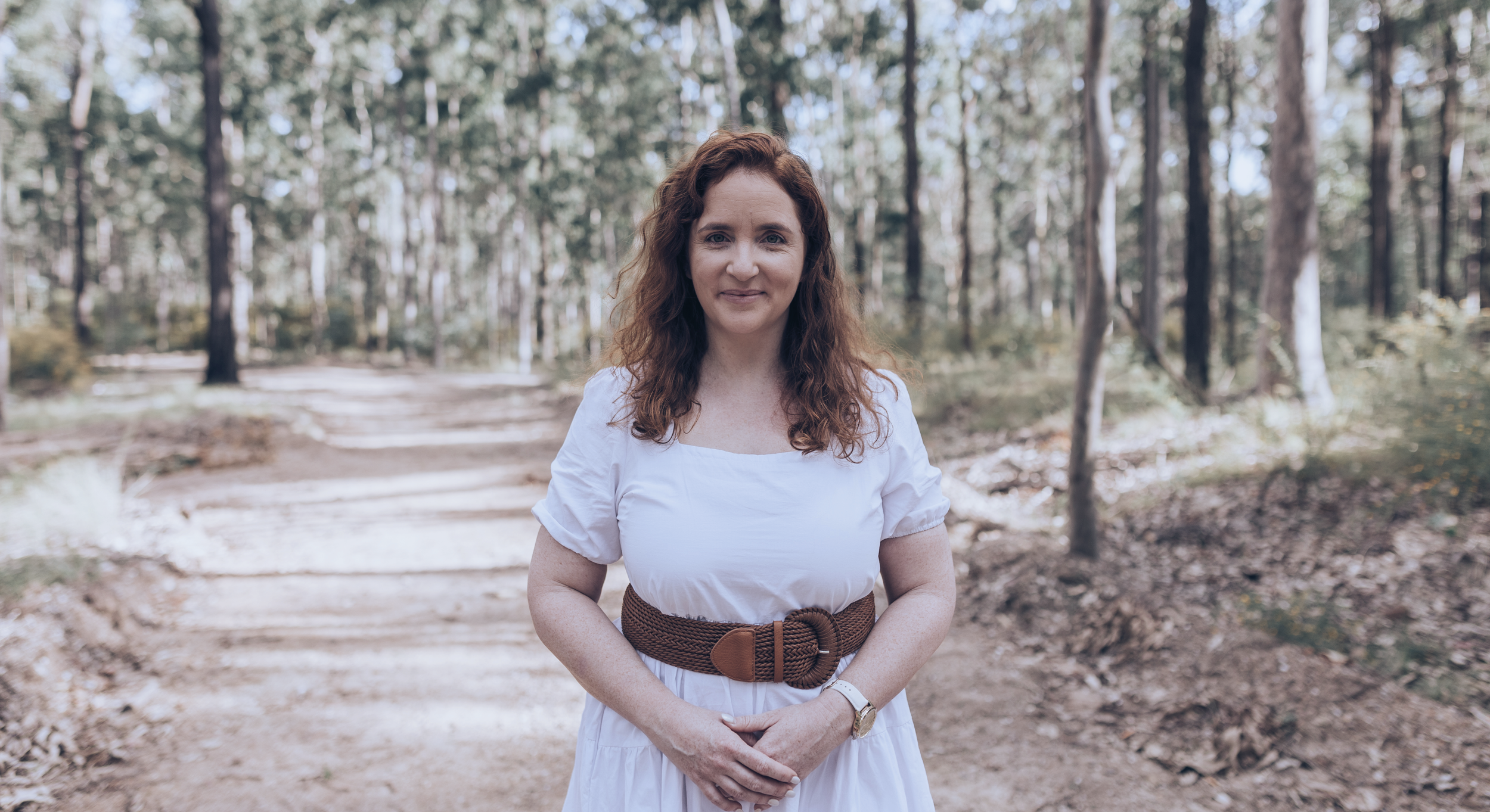 A woman with wavy brown hair wearing a white dress and brown belt standing on a dirt path surrounded by trees in a forest.