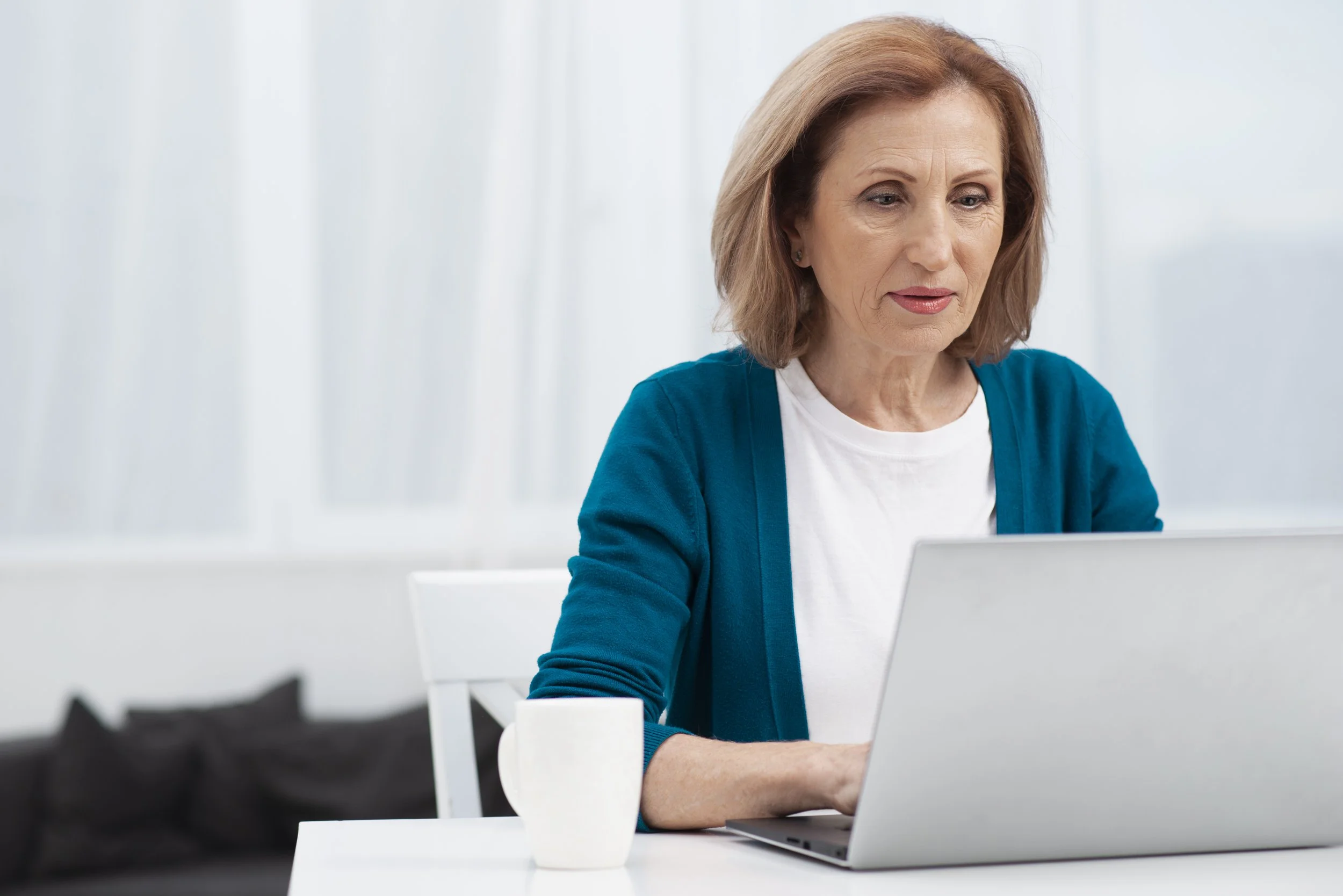 A woman with shoulder-length red hair wearing a white shirt and blue cardigan working on a laptop in a bright, modern office.