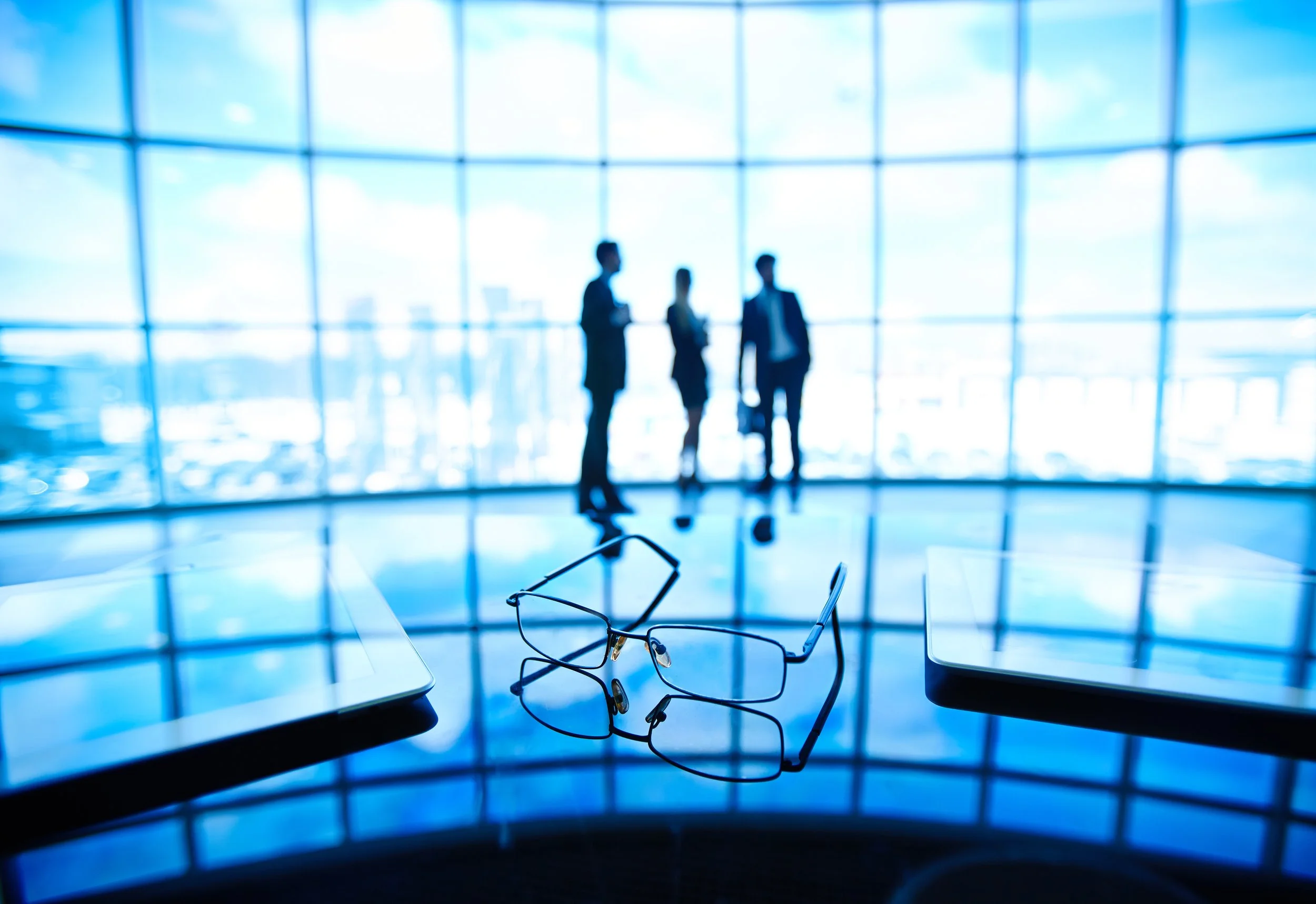 A blurred image of three businesspeople standing and talking inside a modern building with floor-to-ceiling windows showing the city skyline. In the foreground, a pair of eyeglasses is placed on a table.