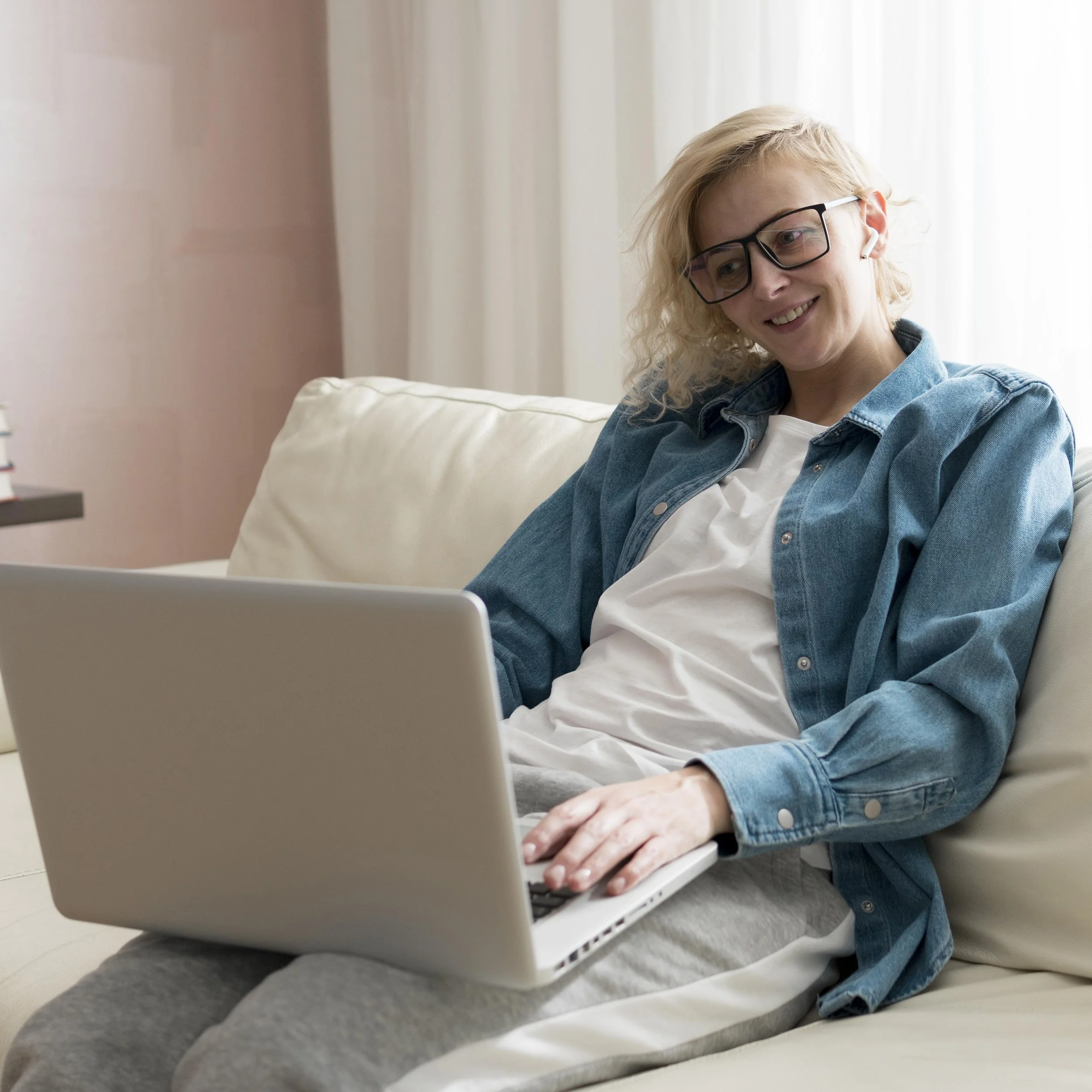 A woman with blonde, curly hair wearing glasses and a denim jacket, sitting on a couch using a laptop.