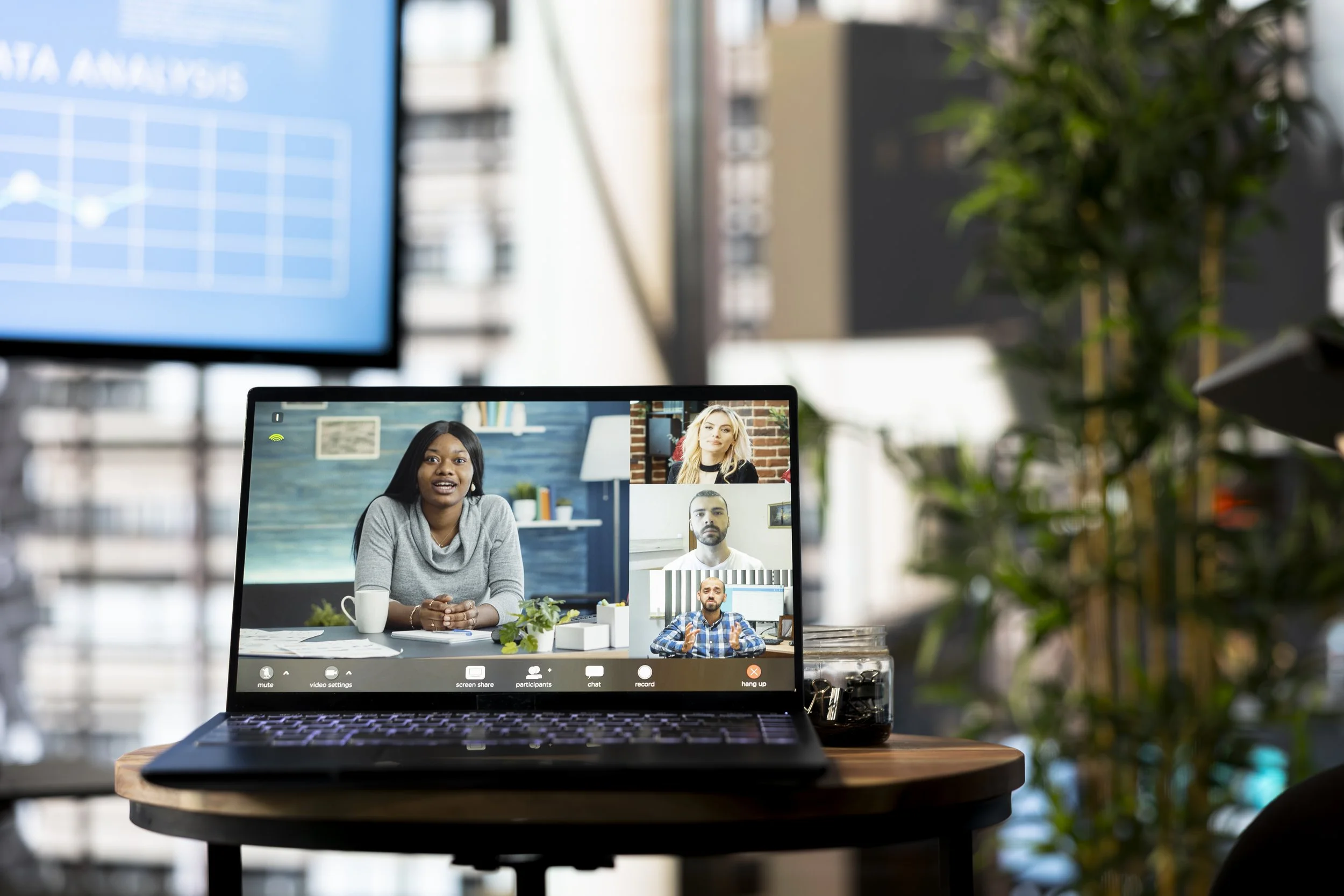 Laptop on a wooden table displays a video conference call with five people, three women and two men, in a bright room with a brick wall and bookshelves. Out of focus, a large window and indoor plants are visible in the background.