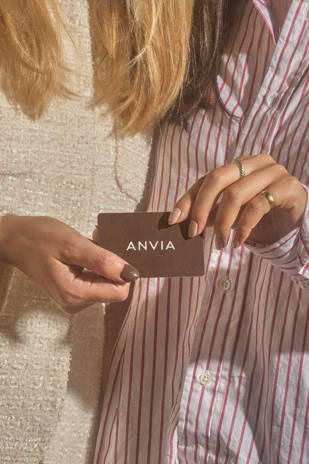 Persona sosteniendo una tarjeta de color marrón con la palabra 'ANVIA' escrita en ella, usando una camisa de rayas blancas y rosas y un suéter crema, con varias uñas pintadas y usando anillos.