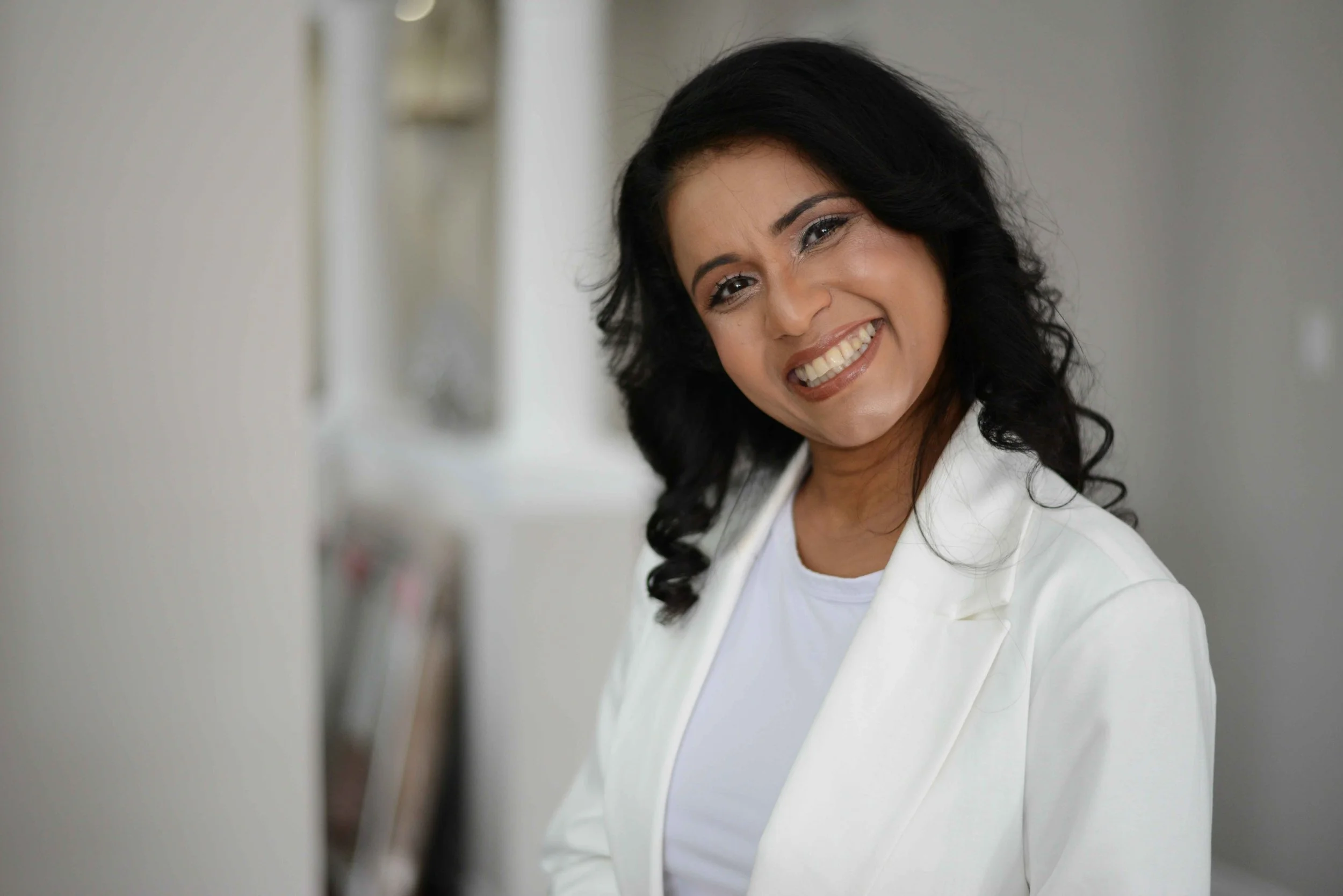 Smiling woman with black wavy hair wearing a white blazer and white shirt, standing indoors near a blurred bookshelf.