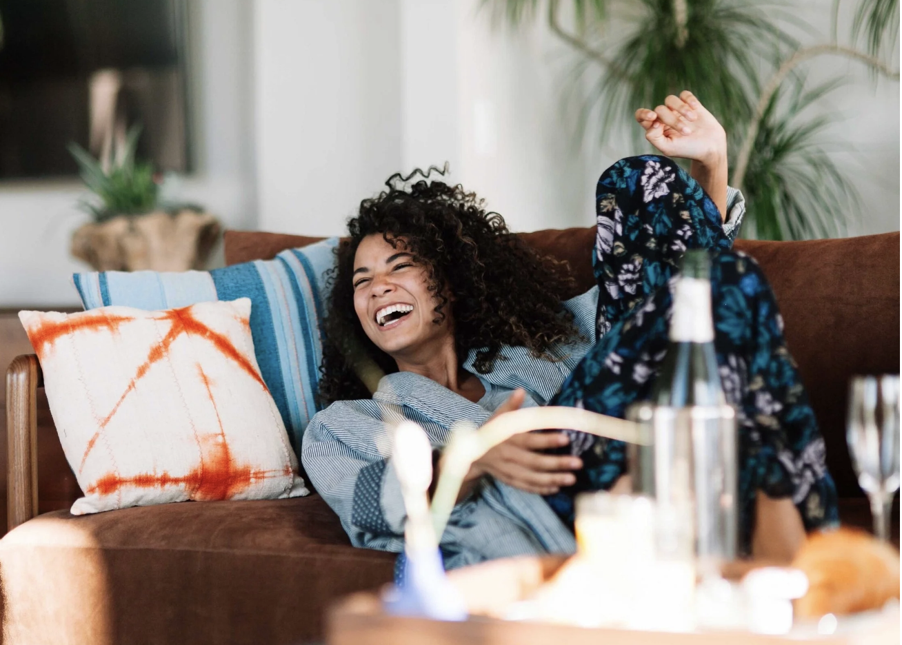 A woman laughing and relaxing on a brown sofa with colorful pillows, a green plant in the background, and a table with drinks and snacks in the foreground.